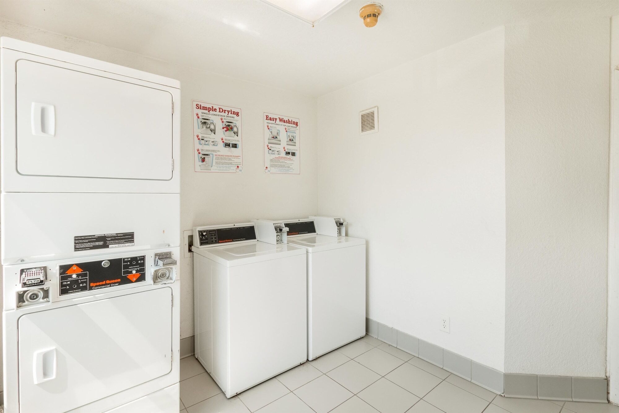 A laundry room with stacked dryers and washing machines, a ceiling light, and two informational posters on the wall.