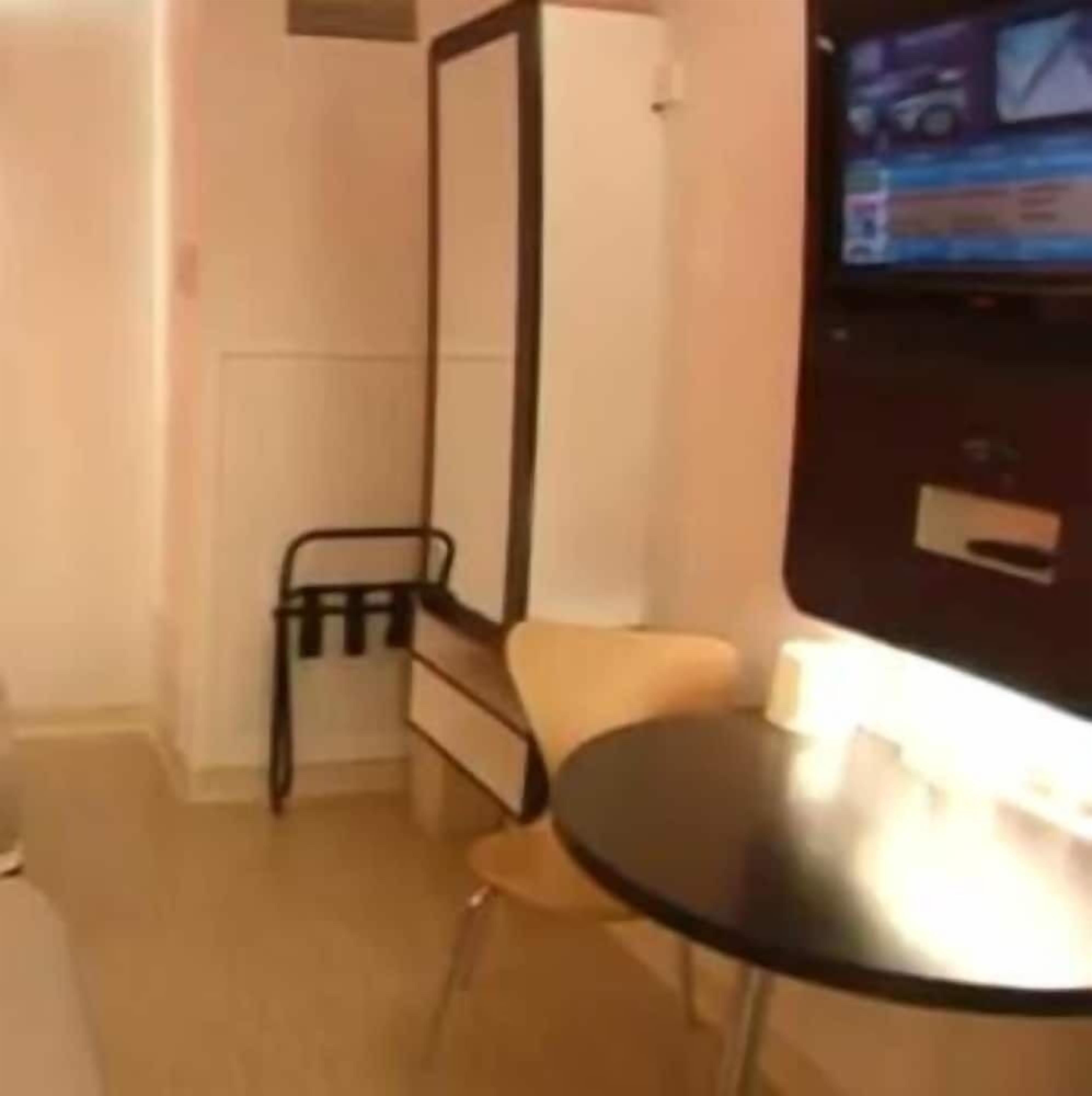 Een kleine kamer met een bed, een stoel, een tafel en een aan de muur bevestigde televisie.