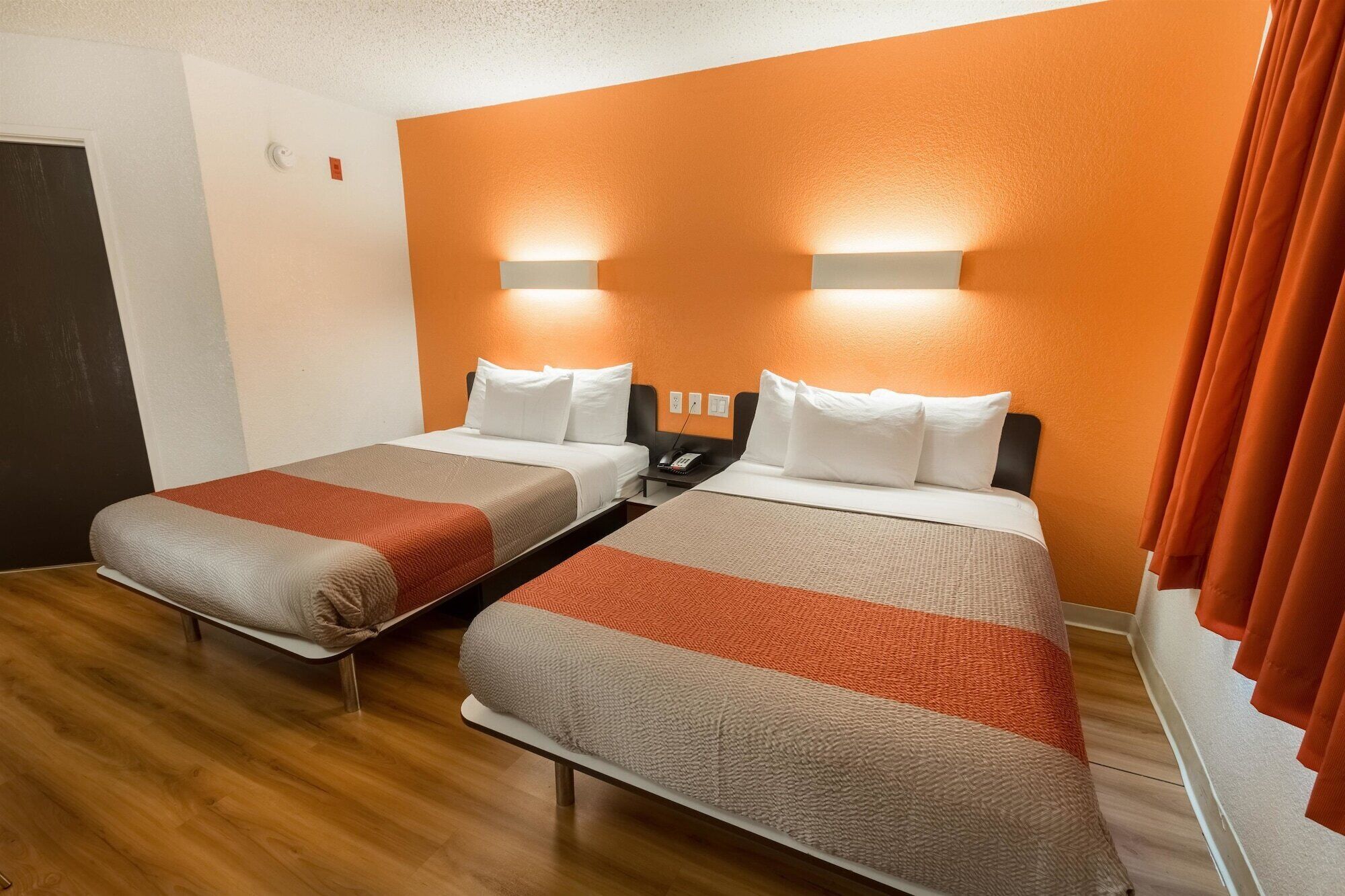 A hotel room with two beds, each with a white headboard and a red and beige blanket. There is a telephone on the nightstand between the beds.