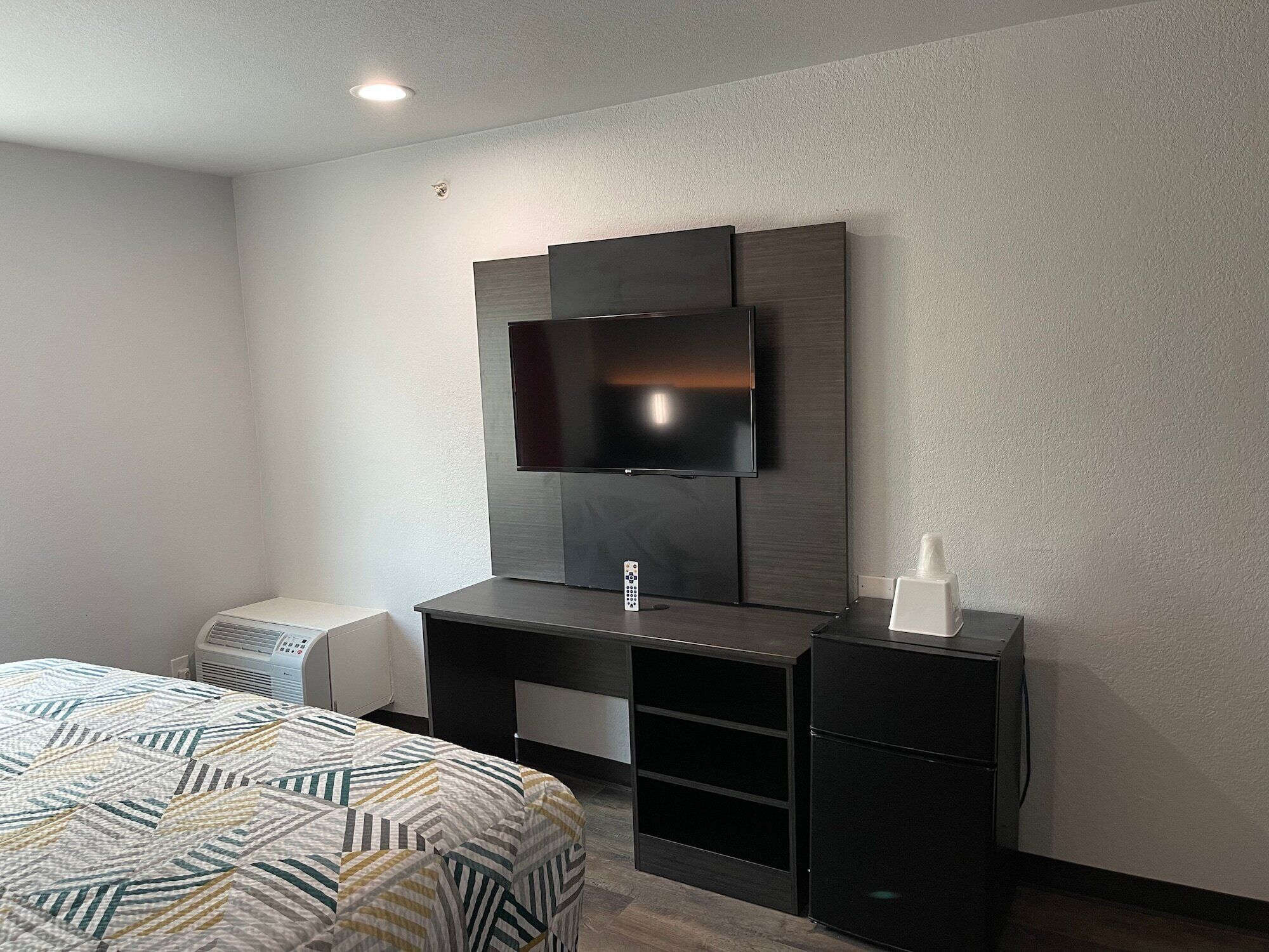 A hotel room with a bed, a desk with a flat-screen TV, a small refrigerator, and a trash bin.