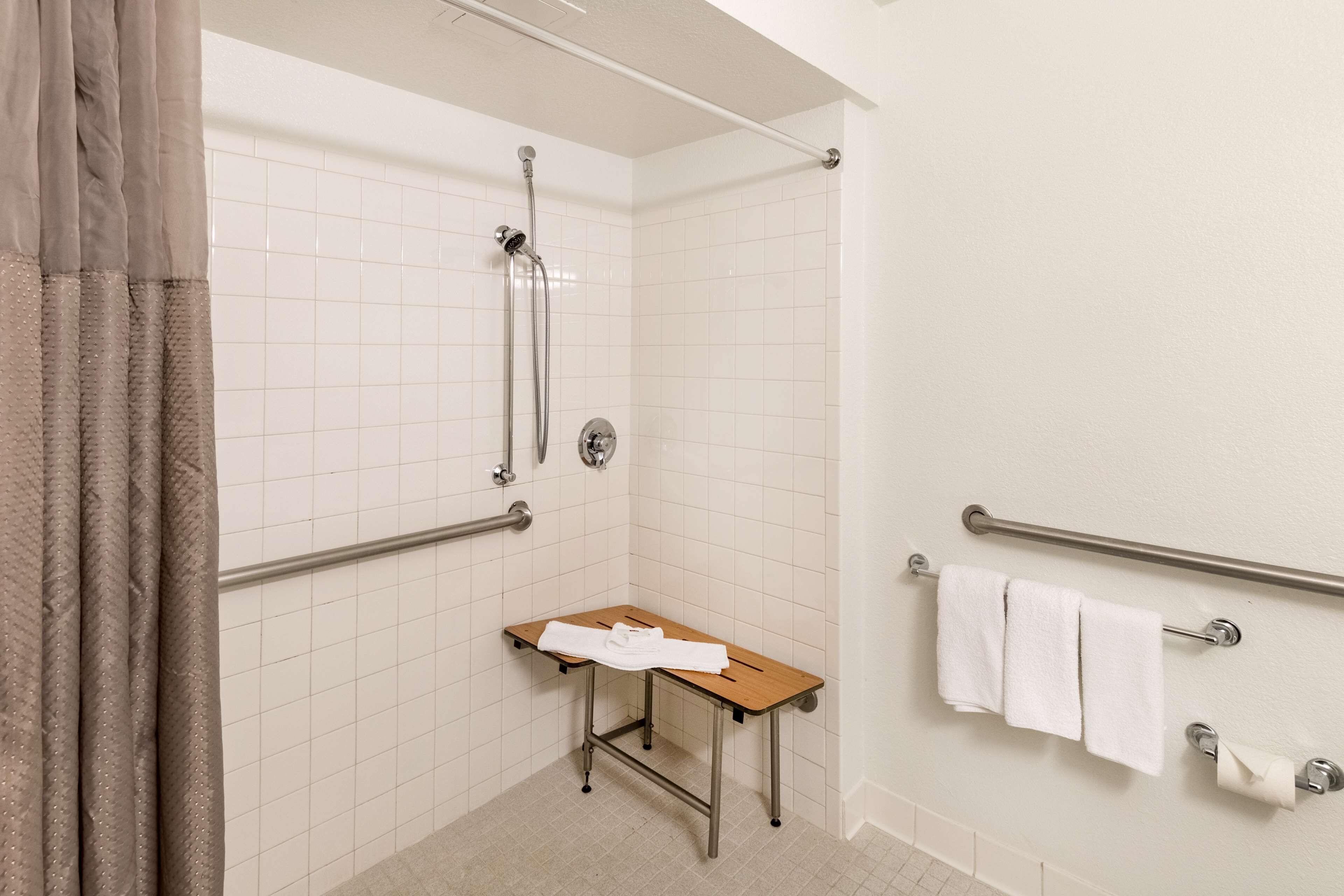 A small bathroom with white tiled walls, a shower, a grab bar, a wooden bench, and a towel rack.