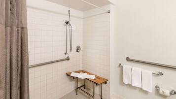 A small bathroom with white tiled walls, a shower, a grab bar, a wooden bench, and a towel rack.