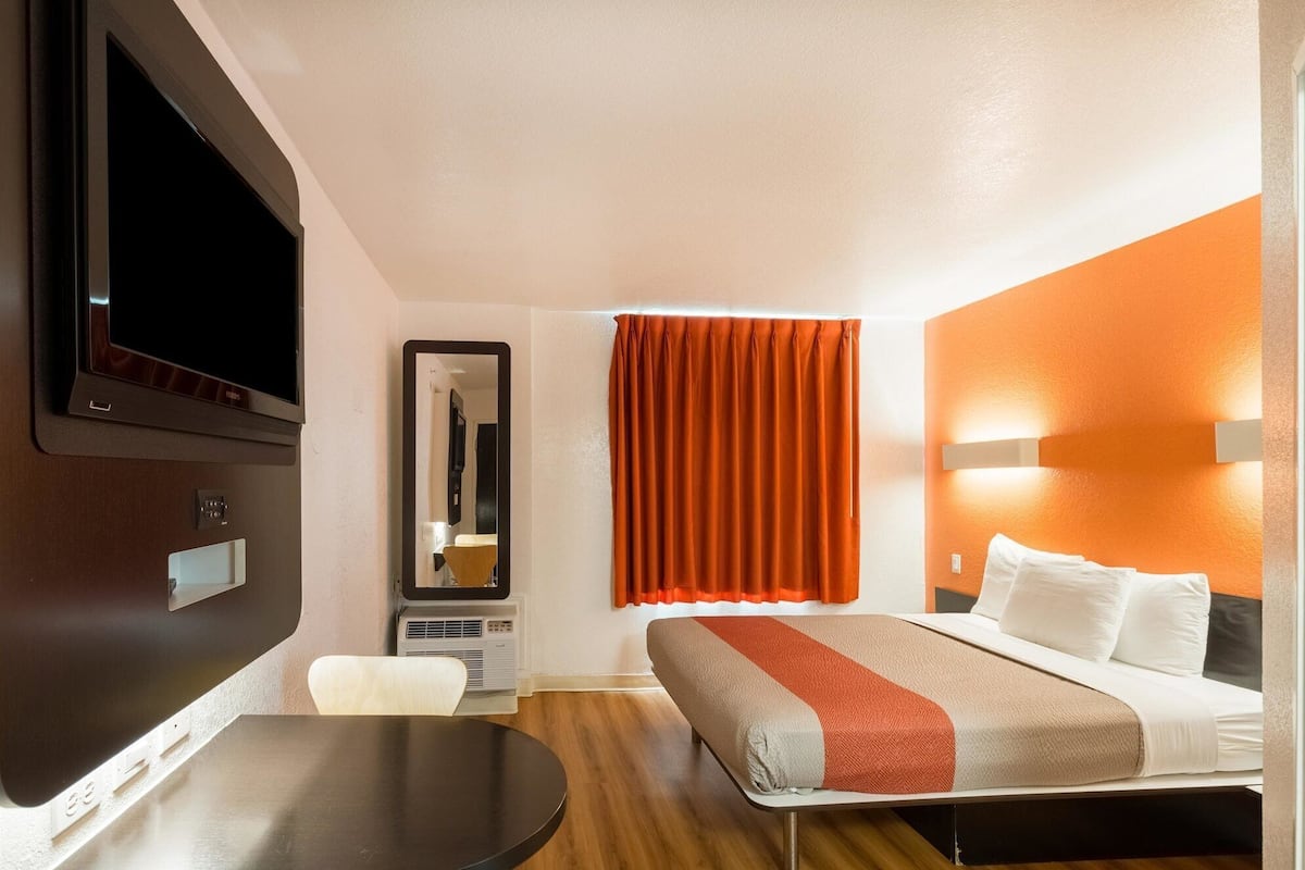 Une chambre d’hôtel avec un lit, un bureau, une chaise, une télévision, un miroir et une fenêtre avec des rideaux orange.