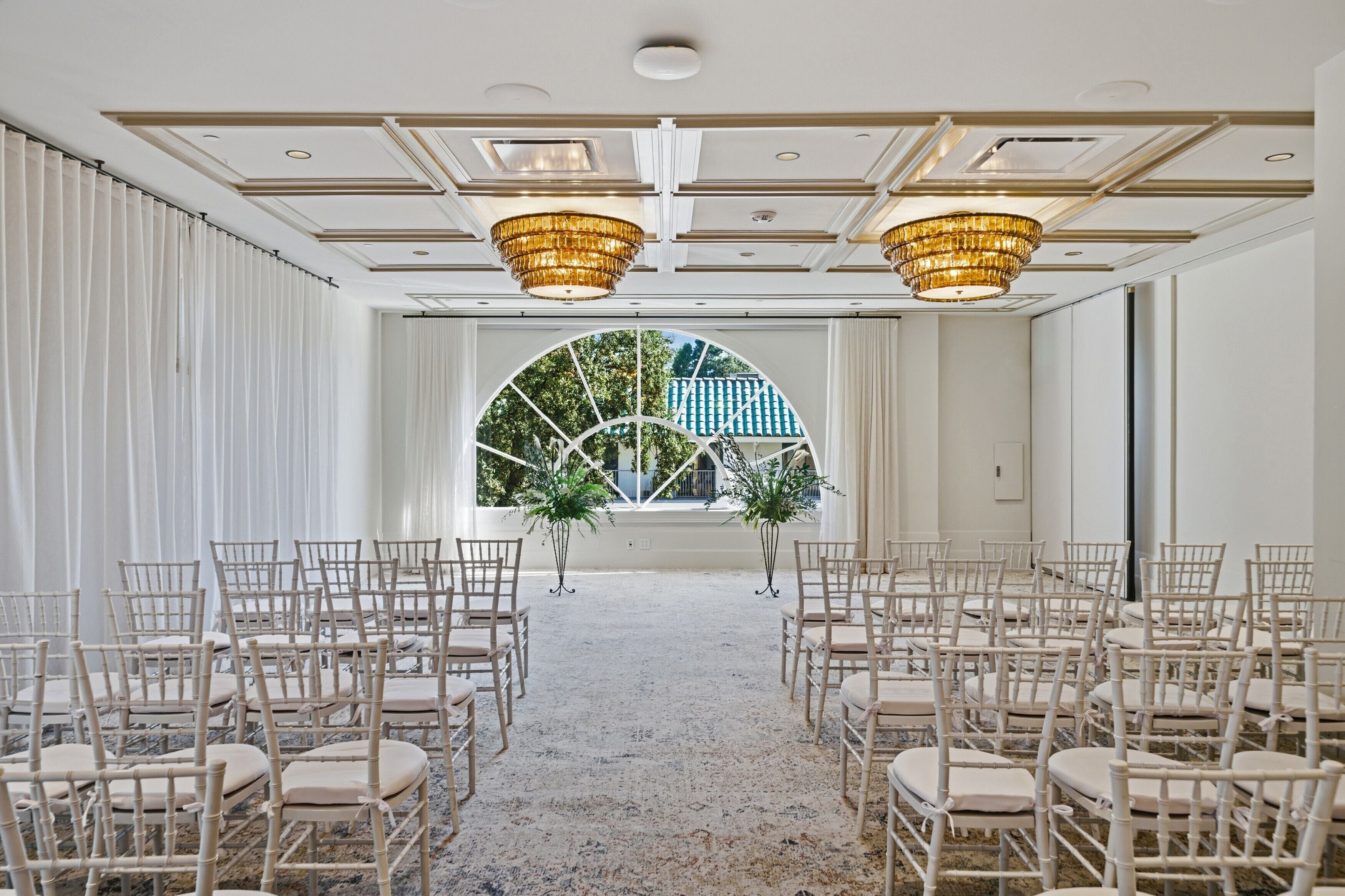 A room with rows of white chairs arranged for an event, a large arched window with a view of greenery, and two chandeliers.