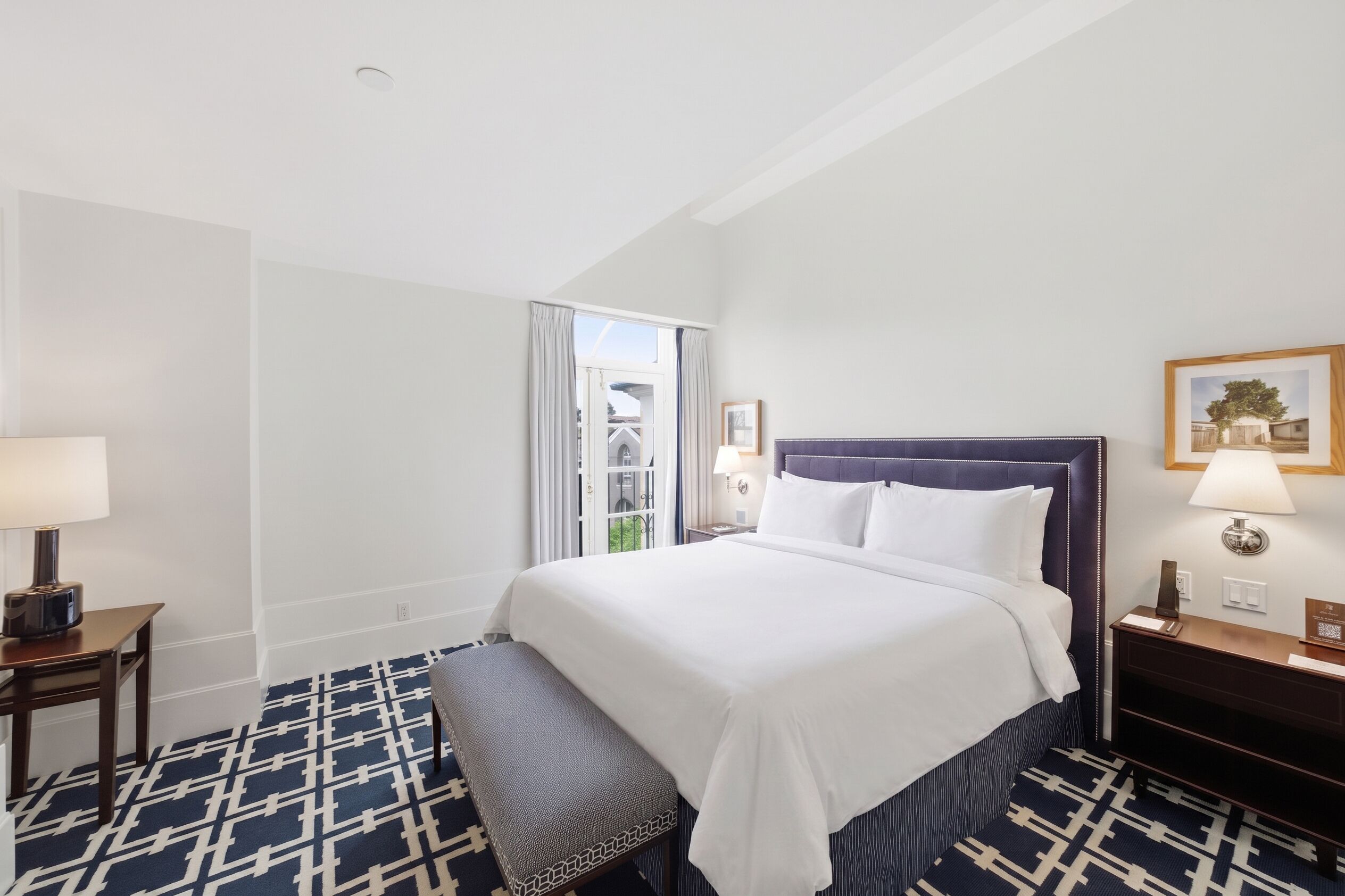 A hotel room with a bed, a bench, a nightstand, a lamp, and a window with curtains.