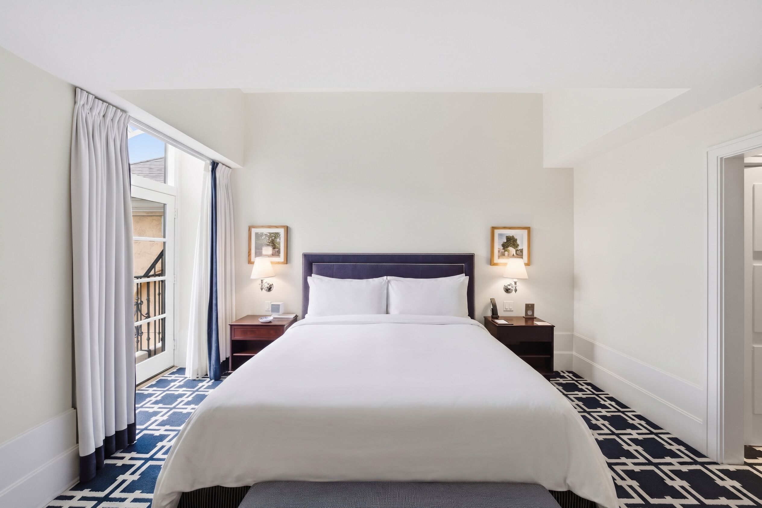 A hotel room with a bed, two bedside tables with lamps, a window with curtains, and a patterned floor.
