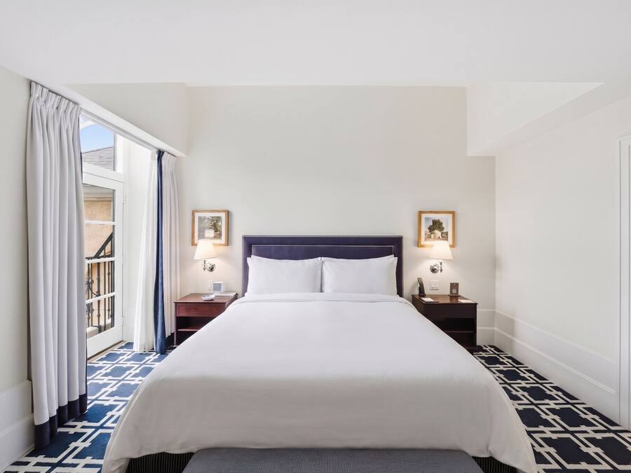 A hotel room with a bed, two bedside tables with lamps, a window with curtains, and a patterned floor.