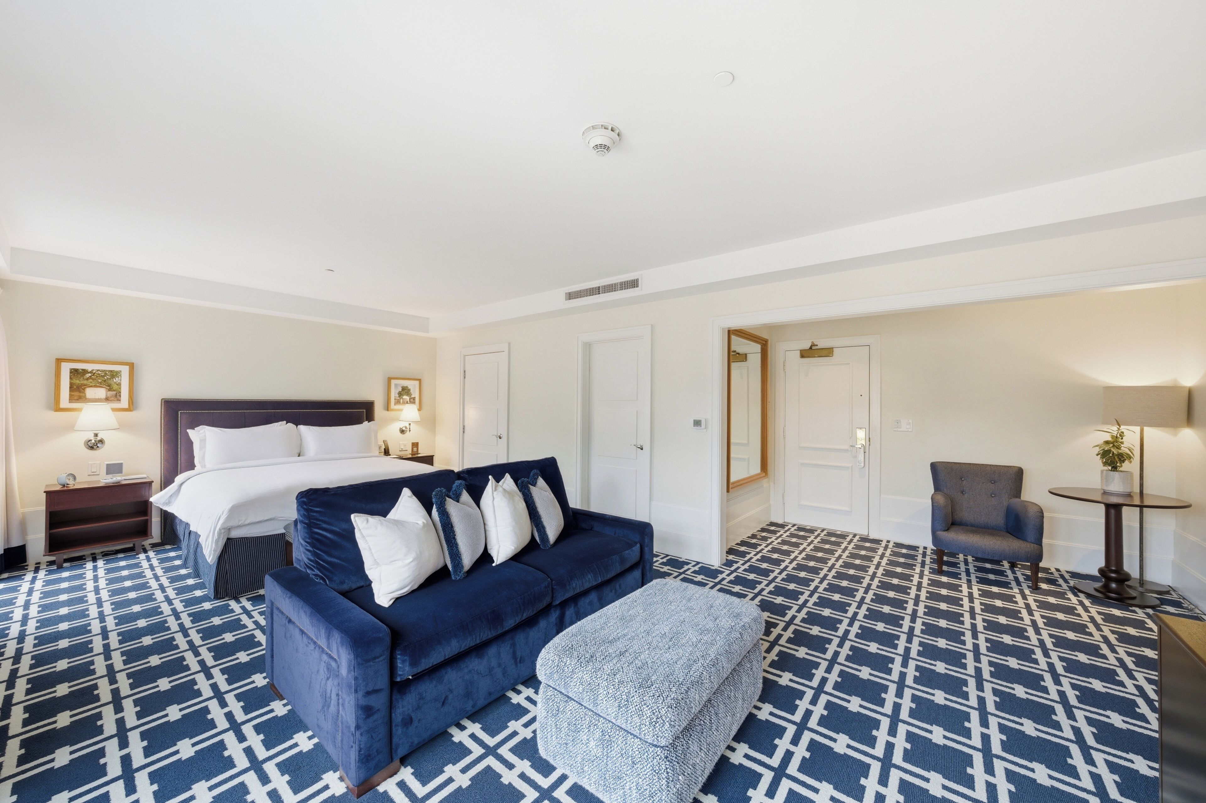 Habitación de hotel con cama, sofá azul, reposapiés, sillón y una mesita con una planta.