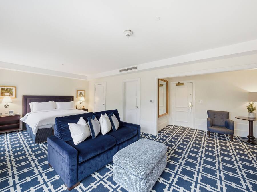 A hotel room with a bed, a blue sofa, an ottoman, a chair, and a small table with a plant.