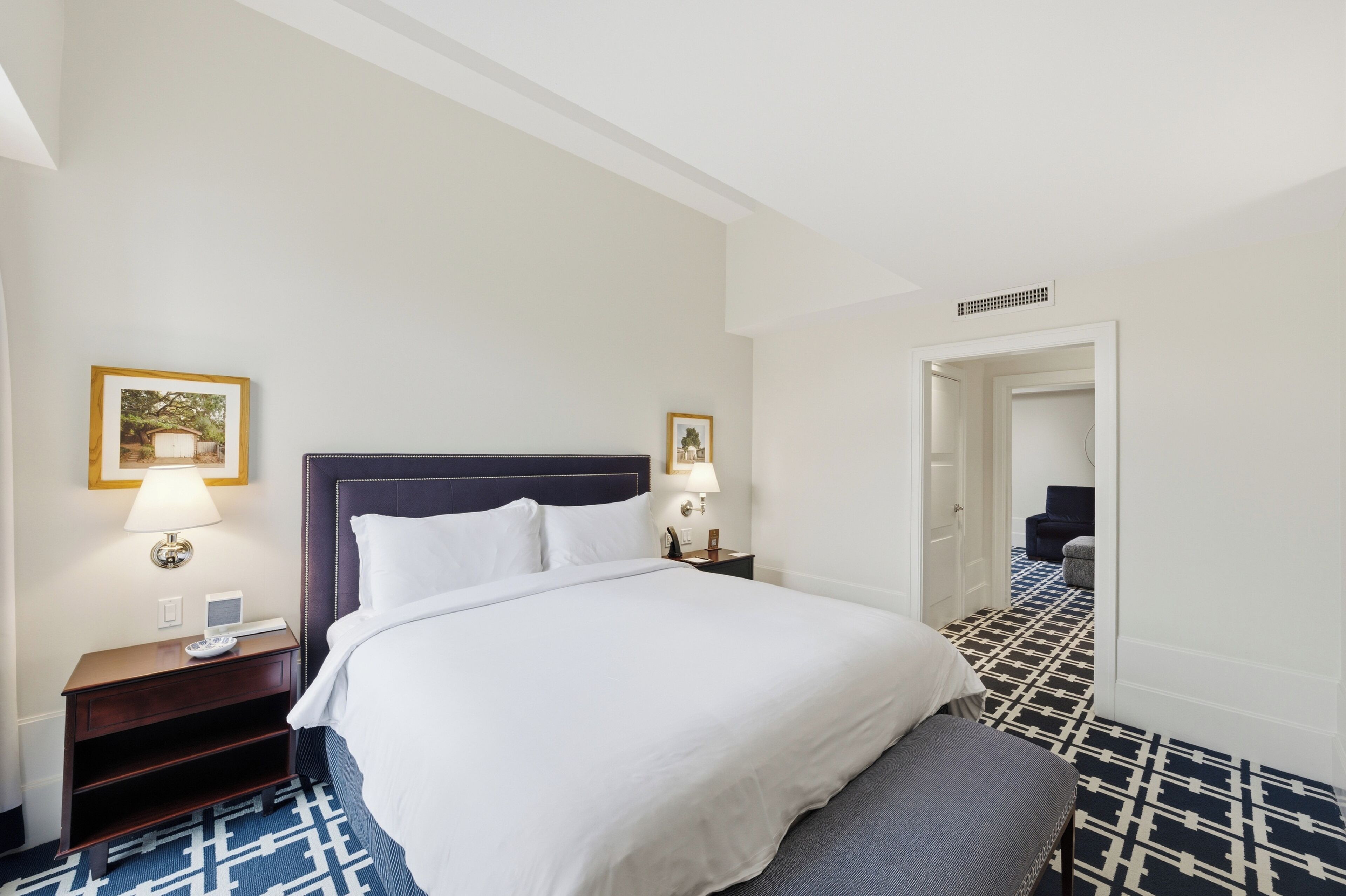 Una habitación de hotel con una cama, mesitas de noche, una lámpara, un marco de cuadros, una silla y una alfombra estampada.