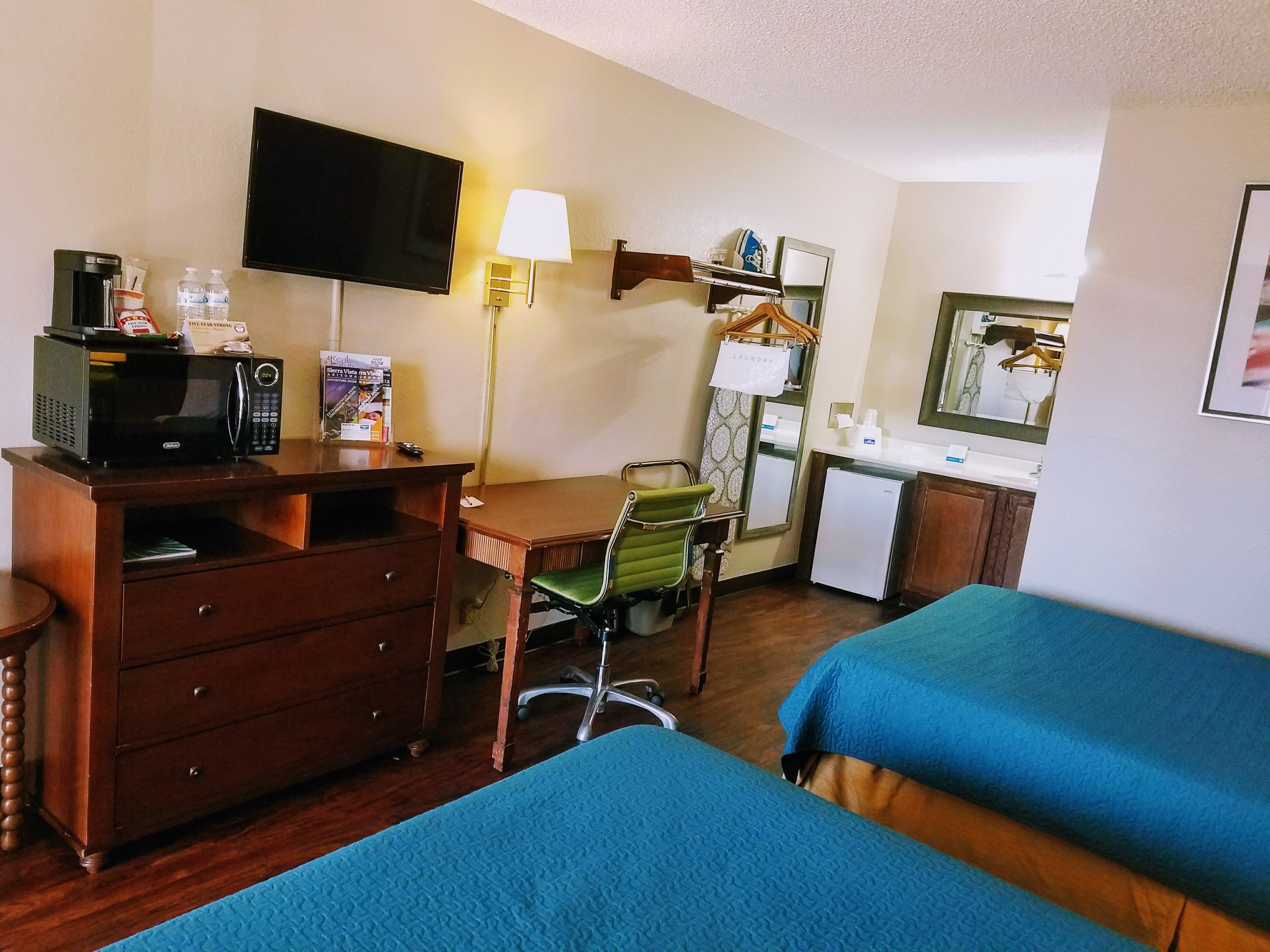Habitación de hotel con escritorio de madera, televisor, microondas, silla, cama con ropa de cama azul, refrigerador pequeño y espejo.
