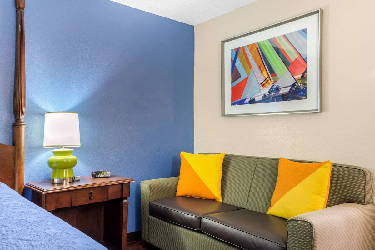 A hotel room with a green sofa, two yellow pillows, a green lamp, and a framed abstract painting on the wall.