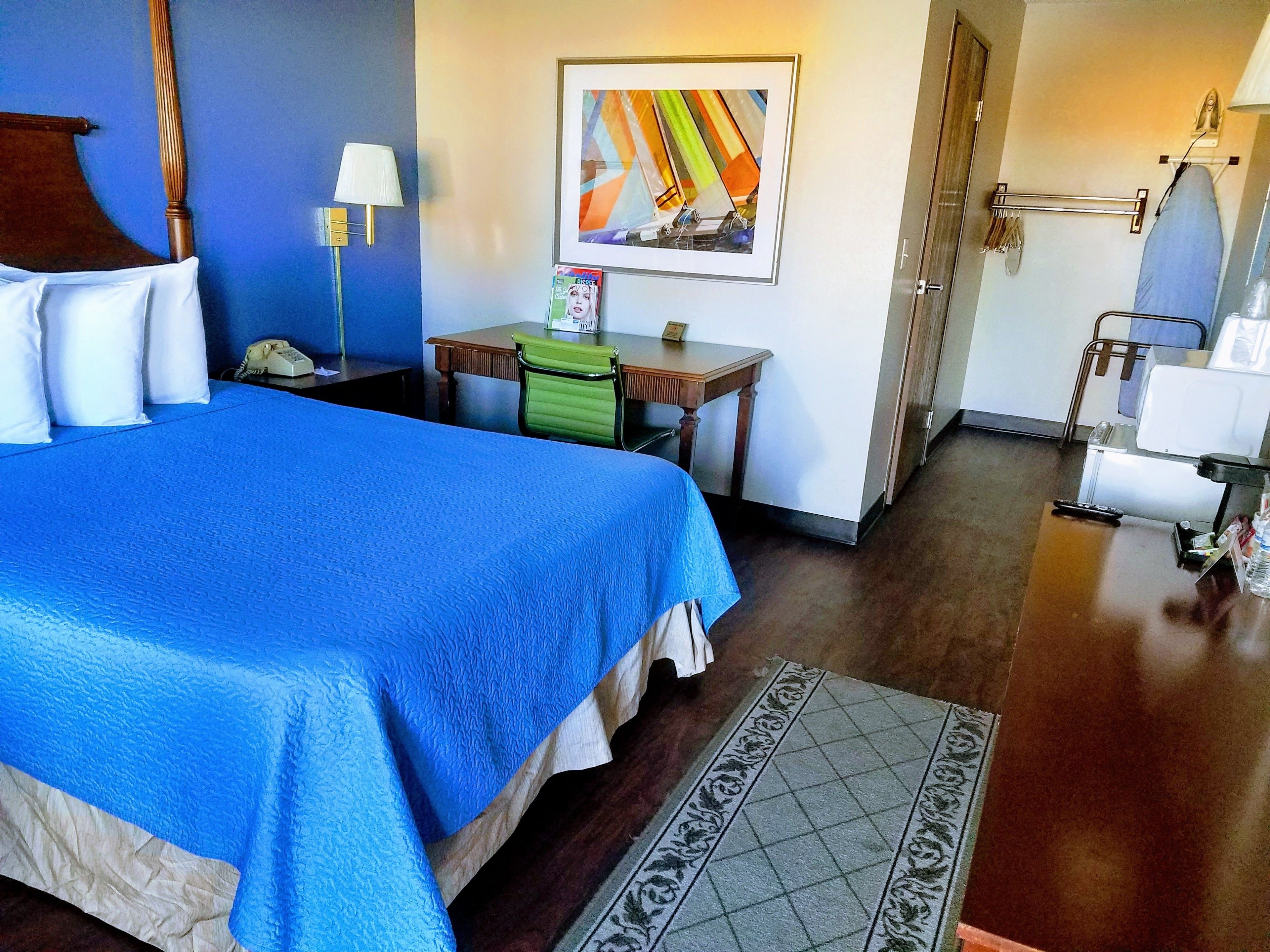 A hotel room with a large bed, a desk, a chair, a painting on the wall, and a bathroom with an iron and towel rack.