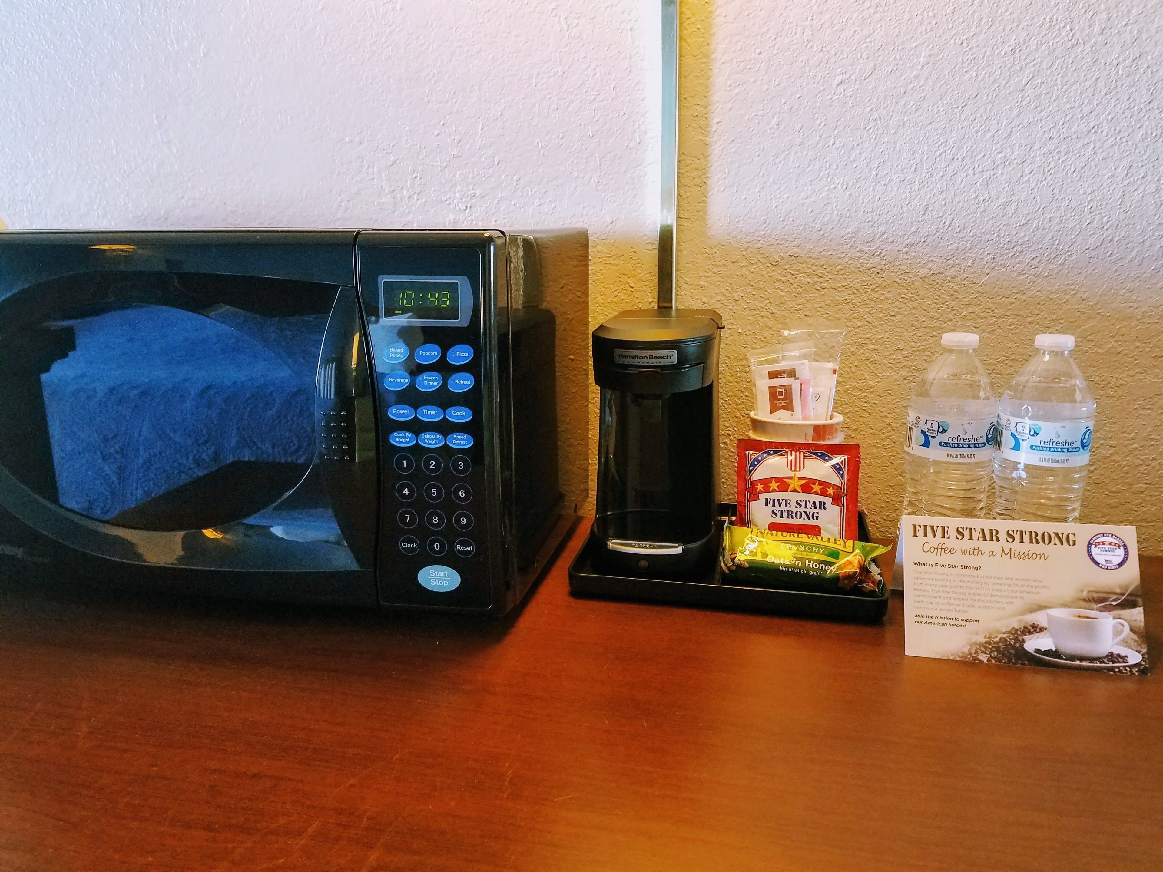A microwave, coffee maker, bottled water, and a coffee bag on a wooden table.