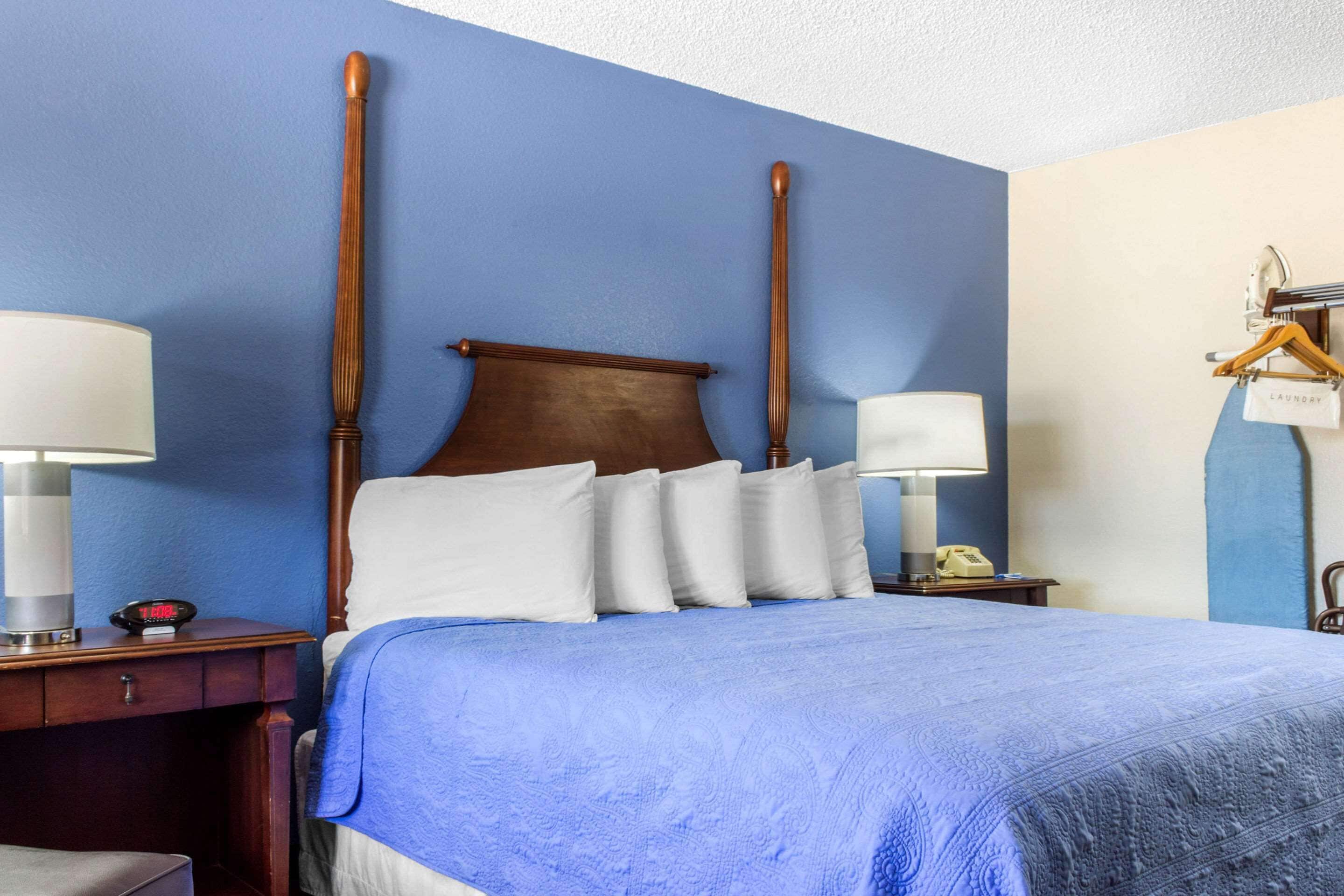 A hotel room with a large bed, two bedside tables, a nightstand, a hanging laundry rack, and a wall clock.