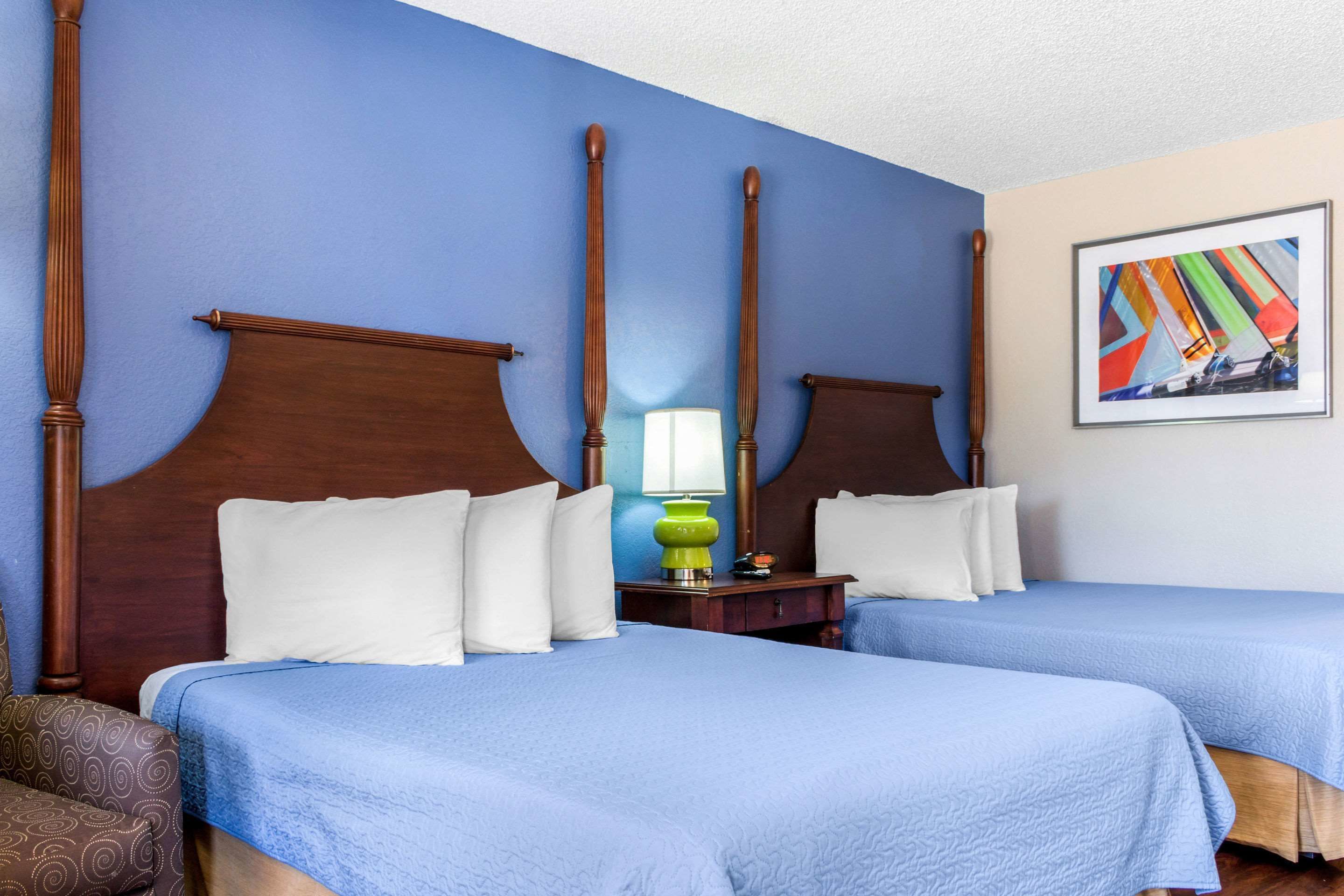 A hotel room with two beds, a wooden headboard, a green lamp, and a colorful painting on the wall.