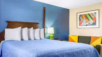 A hotel room with a large bed, a wooden headboard, a green nightstand, a lamp, a colorful painting, and a blue wall.