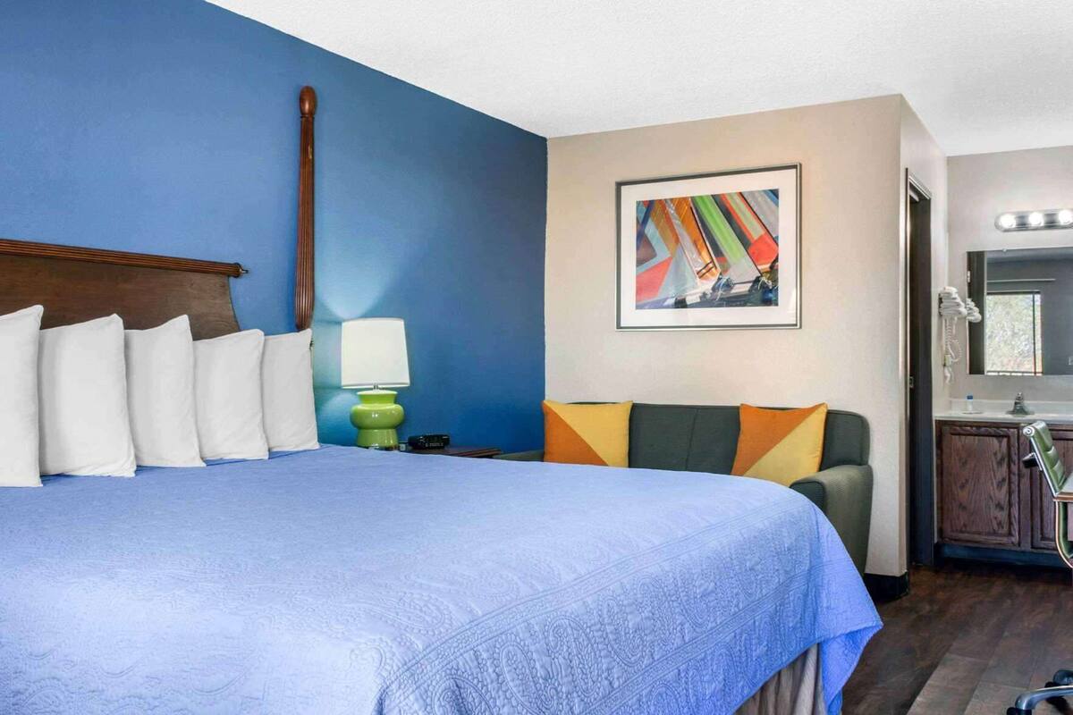 Une chambre d’hôtel avec un grand lit, un bureau, une chaise, une lampe, un tableau et une salle de bain avec un miroir et un lavabo.
