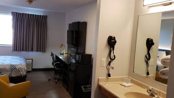 A hotel room with a bed, desk, chair, TV, hairdryer, and a bathroom with a sink and mirror.