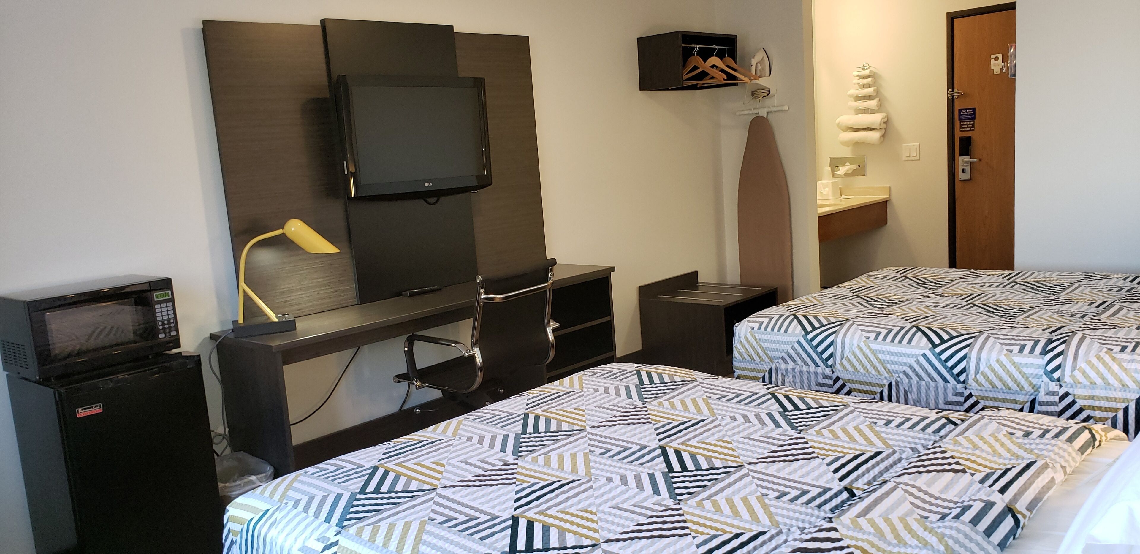 A hotel room with a TV mounted on the wall, a desk with a lamp, a microwave, and two beds with patterned bedding.