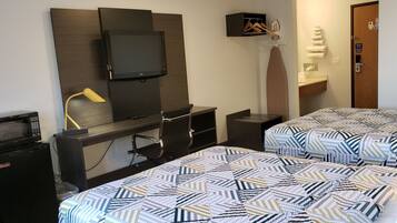 A hotel room with a TV mounted on the wall, a desk with a lamp, a microwave, and two beds with patterned bedding.