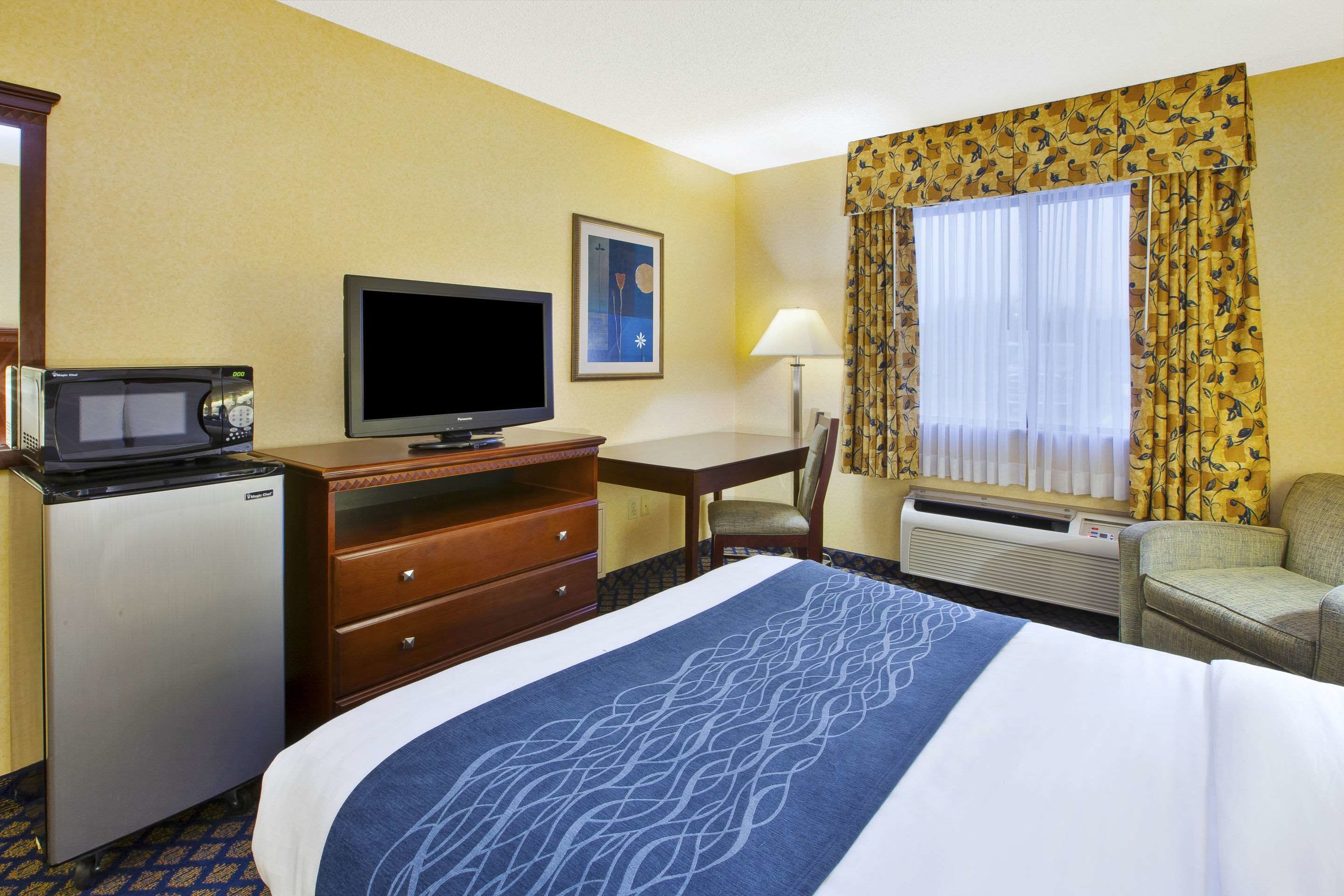 A hotel room with a bed, a desk with a computer, a television, a microwave, a small refrigerator, a chair, a window with curtains, and an air conditioning unit.