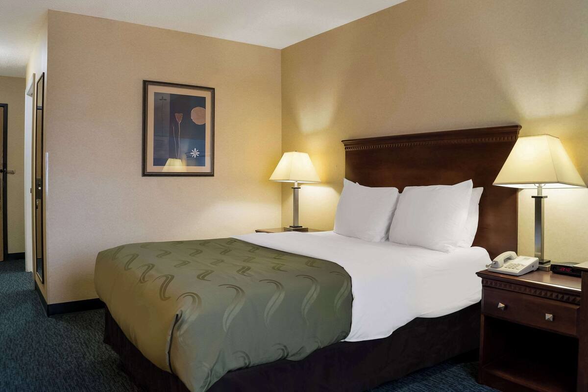 A hotel room with a large bed, two bedside lamps, a bedside phone, a framed picture on the wall, and a door to the left.