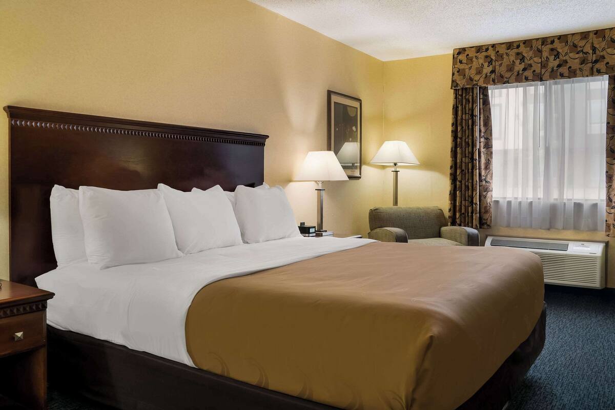 A hotel room with a large bed, two bedside lamps, a chair, a window with curtains, and an air conditioning unit.