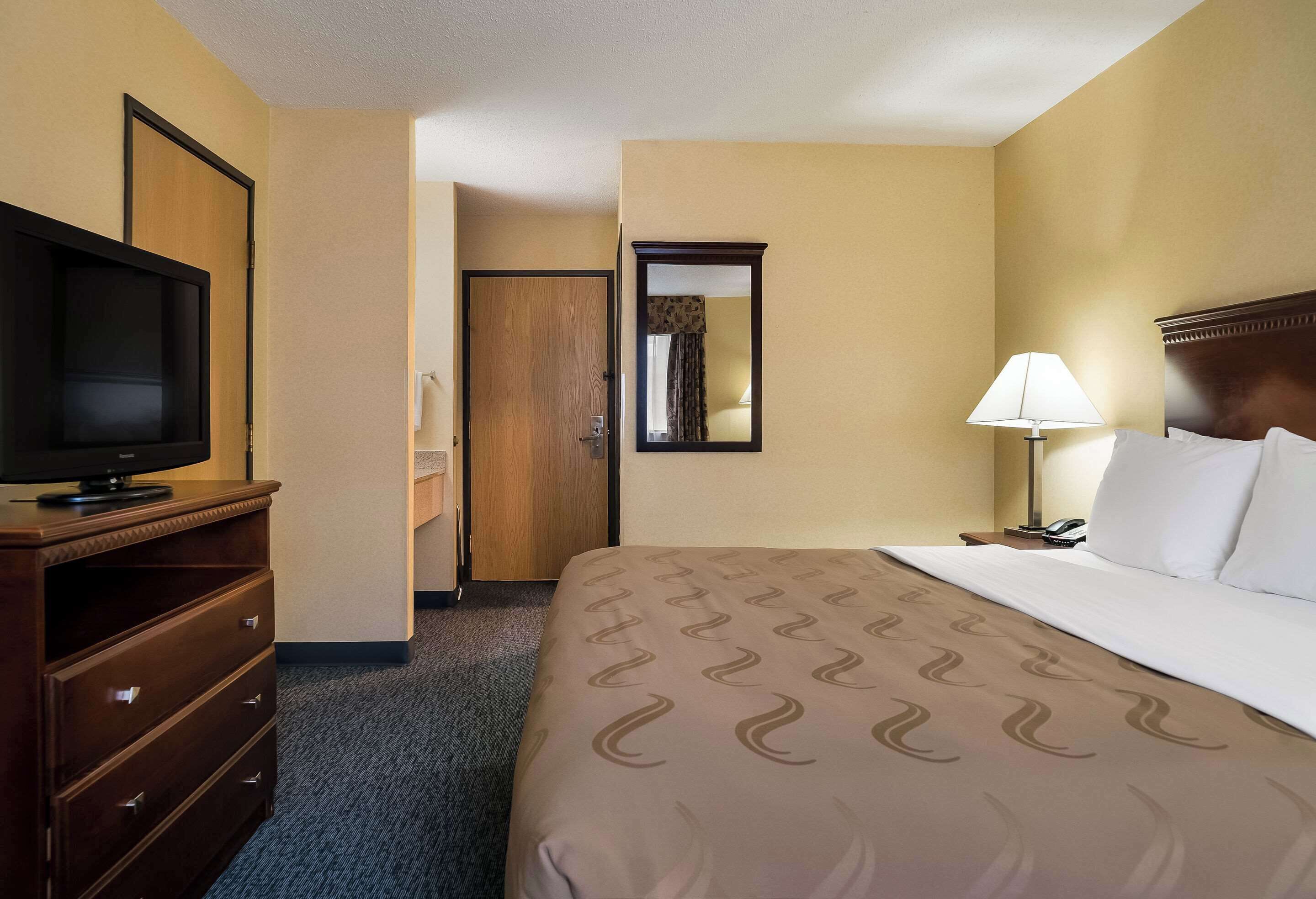 A hotel room with a bed, a TV on a wooden dresser, a lamp, and a door leading to another room.