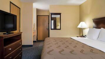 A hotel room with a bed, a TV on a wooden dresser, a lamp, and a door leading to another room.