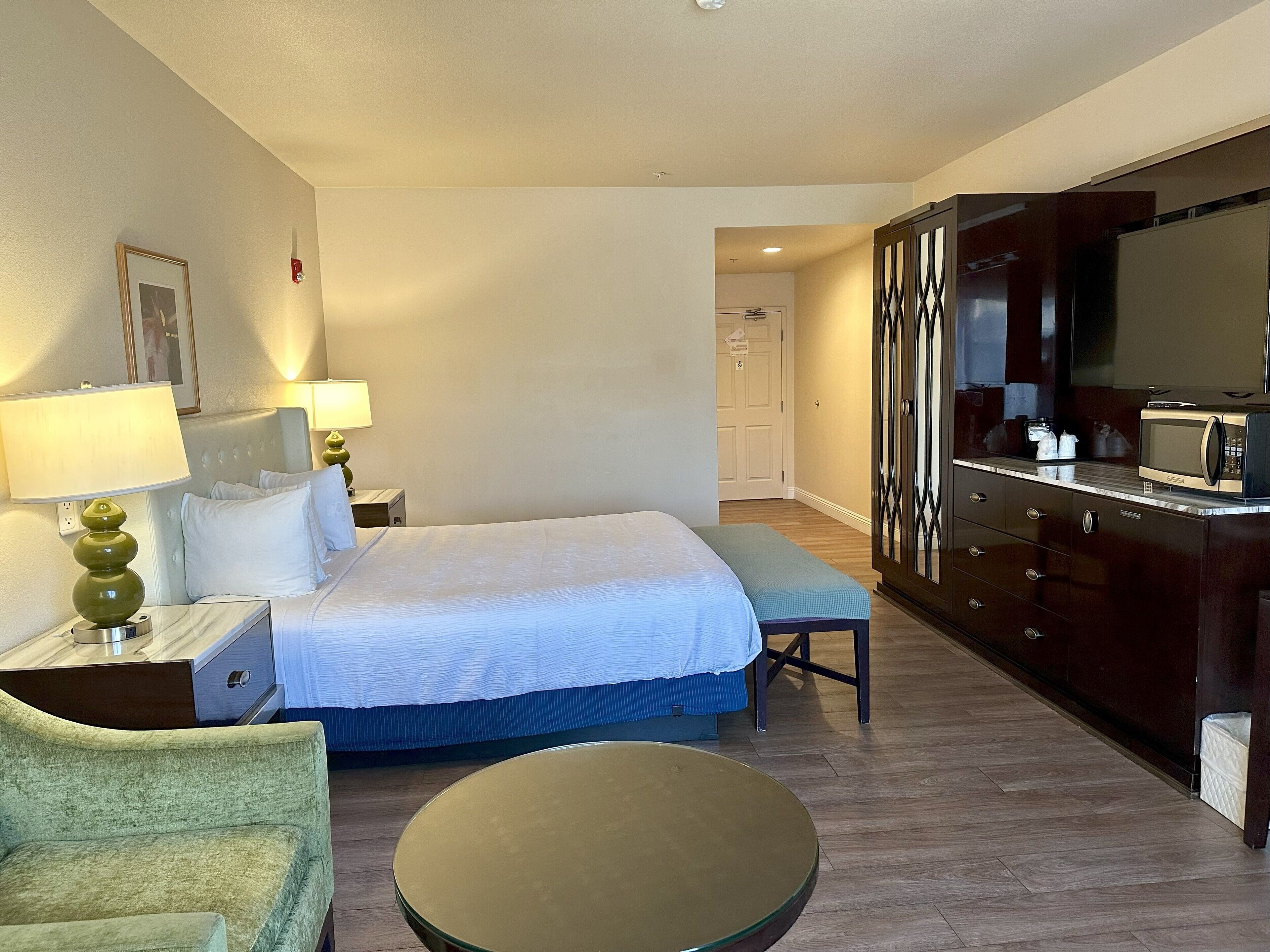 A hotel room with a bed, bedside tables, a chair, a small table, a dresser, and a TV.