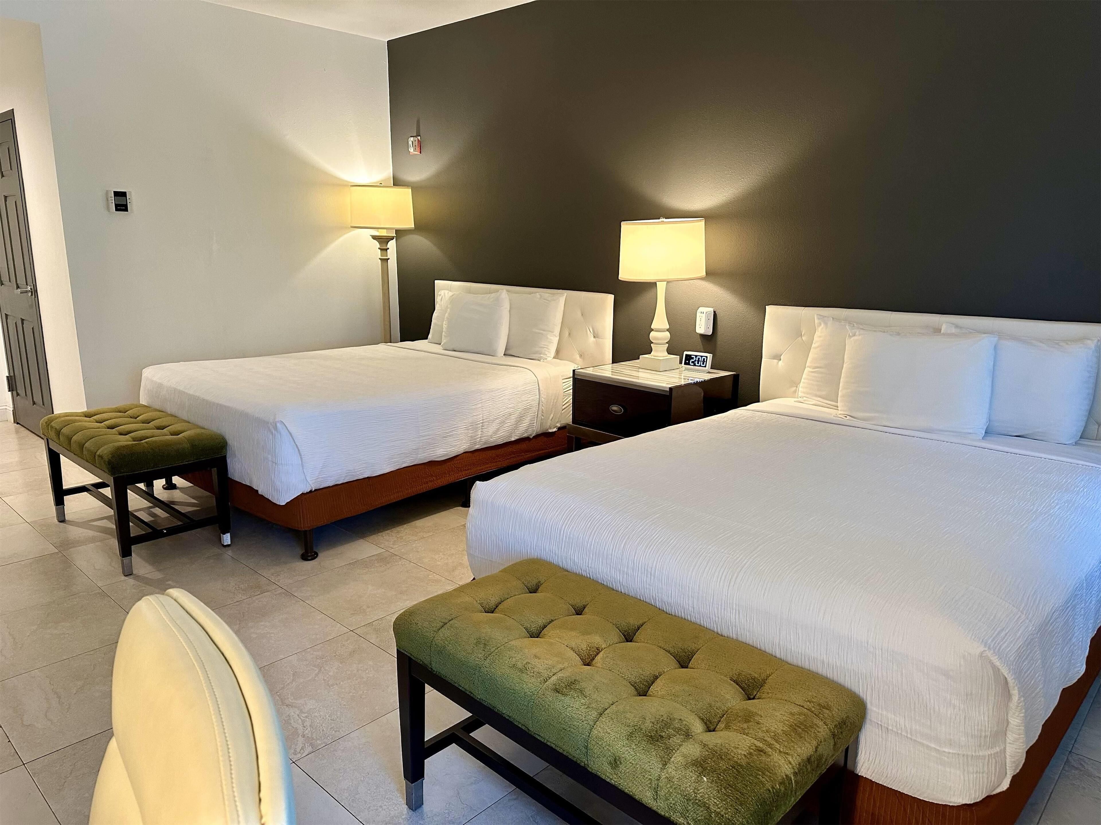 A hotel room with two beds, a green ottoman, a nightstand with a lamp, and a chair.