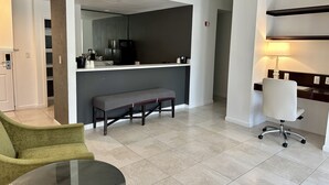 A modern living room with a glass coffee table, a green armchair, a grey bench, and a white office chair. There is a kitchen area with a microwave and a coffee machine. - Rocklin Park Hotel, Trademark Collection by Wyndham (Rocklin)
