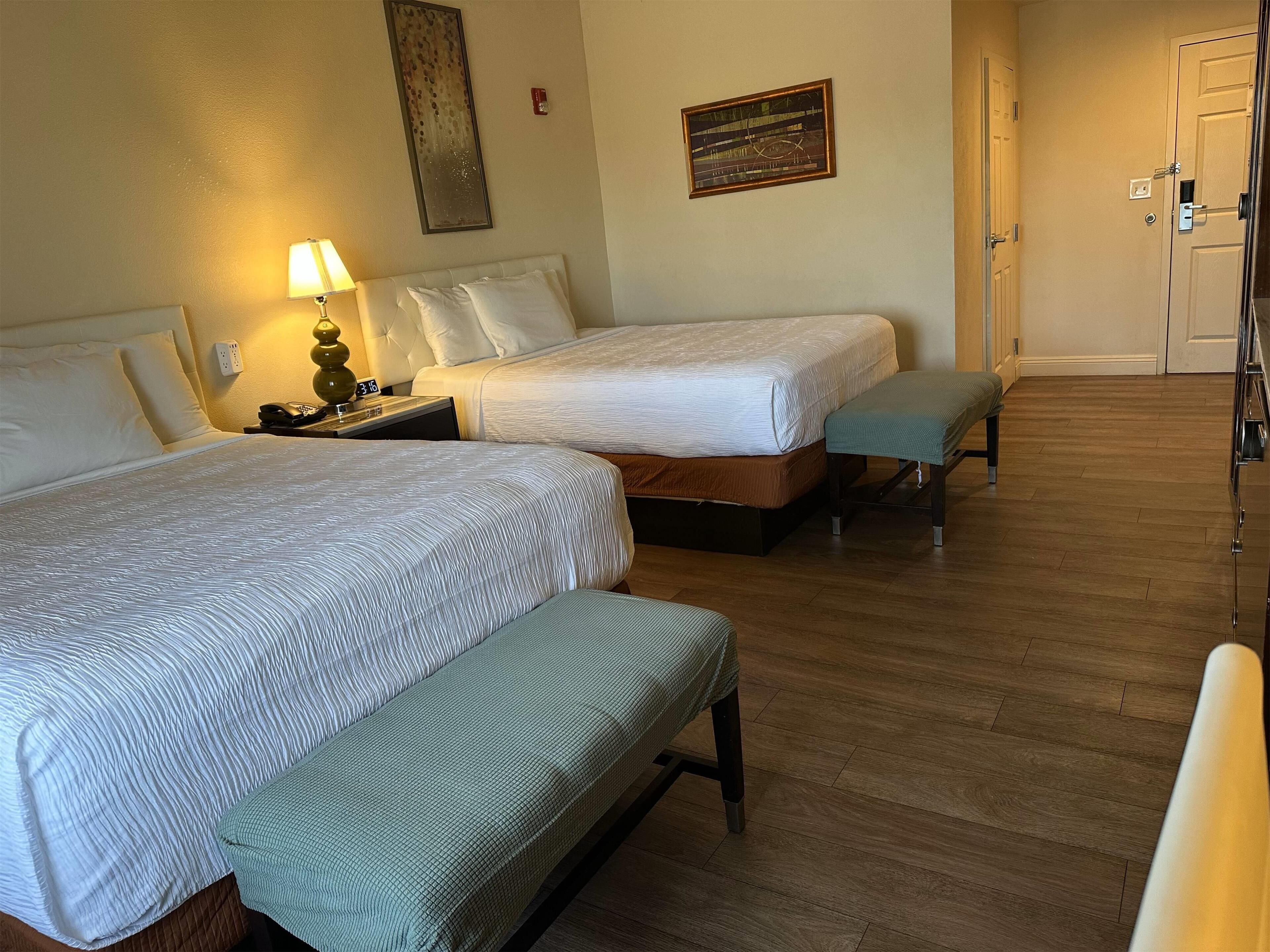 Una habitación de hotel con dos camas, un banco, una mesita de noche con lámpara y una puerta.