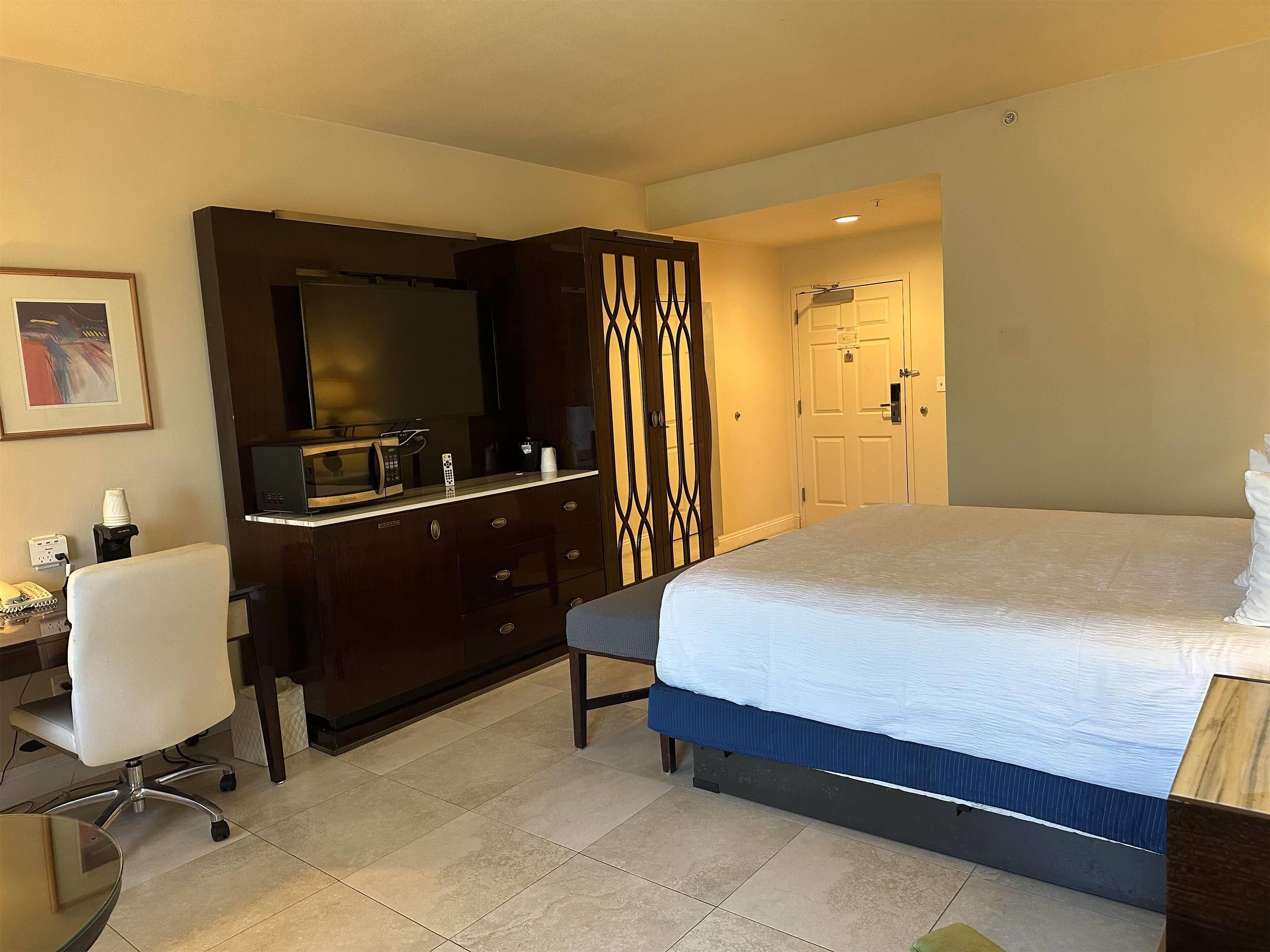 A hotel room with a bed, a desk with a chair, a wardrobe, a mirror, and a door.