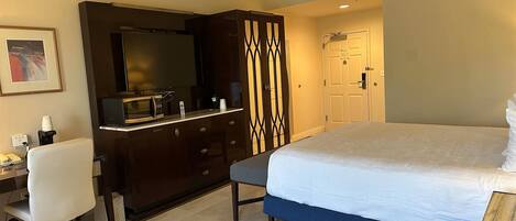 A hotel room with a bed, a desk with a chair, a wardrobe, a mirror, and a door.