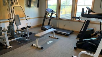 Uma academia com equipamentos de ginástica, um banco, esteiras e uma TV fixada na parede.
