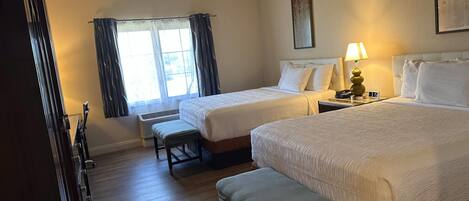 A hotel room with two beds, a bench, a window with curtains, a wall clock, and a lamp.