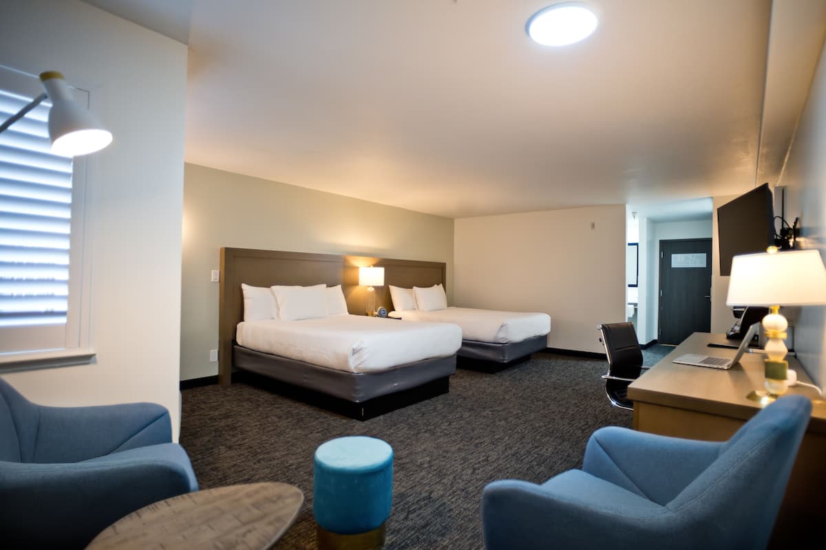 A hotel room with two beds, a desk with a laptop, a chair, a small table, and a lamp.