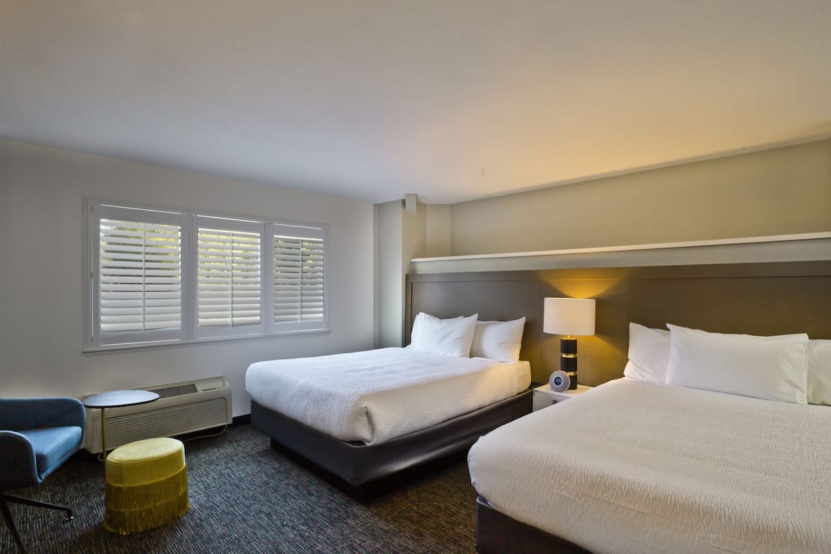 A hotel room with two beds, a desk, a chair, and a lamp.