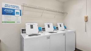 A laundromat with four washing machines, a phone, and a poster advertising a laundry app. - Extended Stay America Suites Dallas Plano Parkway (Plano)