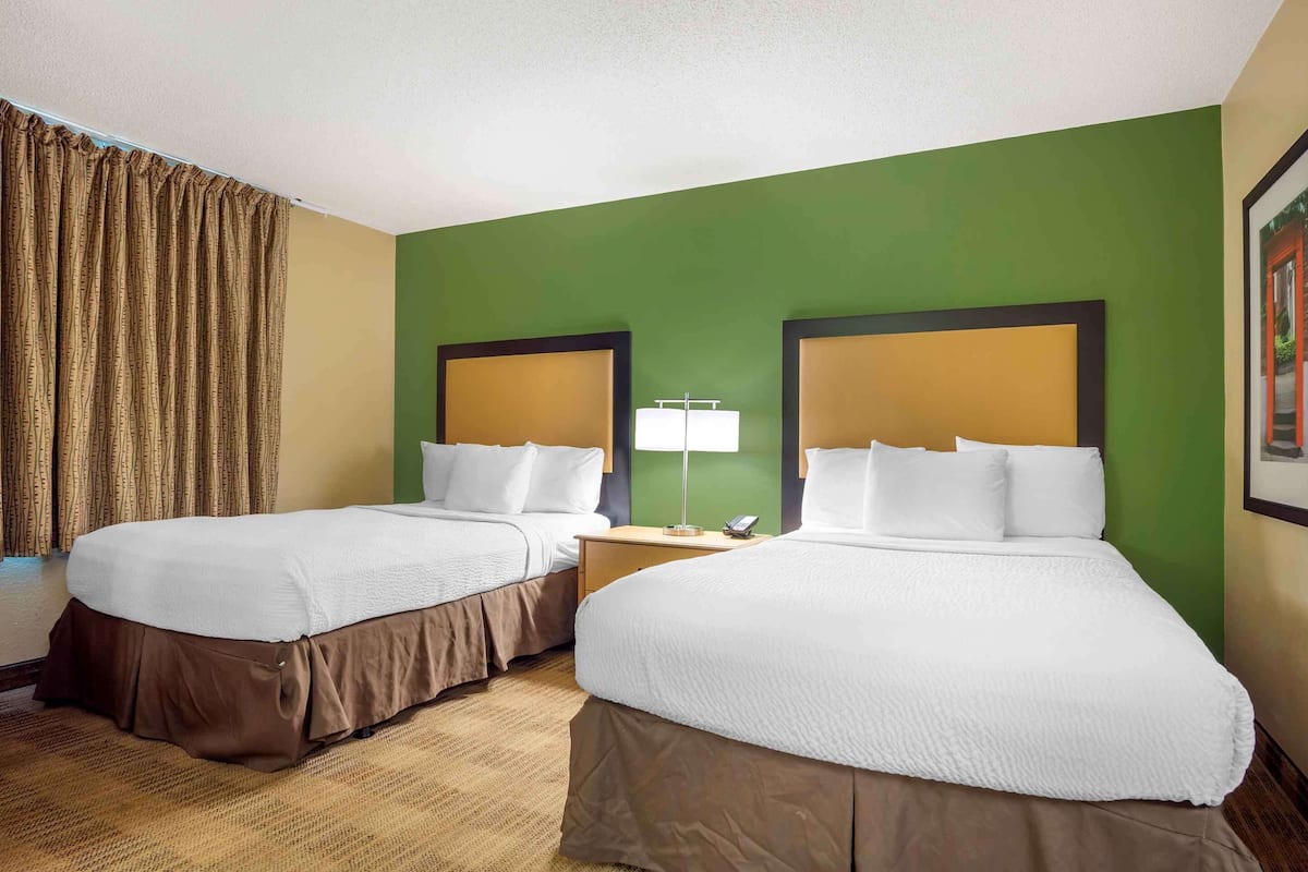 A hotel room with two beds, a nightstand, a lamp, and a window with curtains.