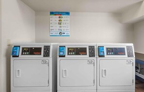 Three commercial dryers with digital displays and control panels, mounted on a wall with a laundry tips poster above them. - Extended Stay America Suites Dallas Plano Parkway (Plano)