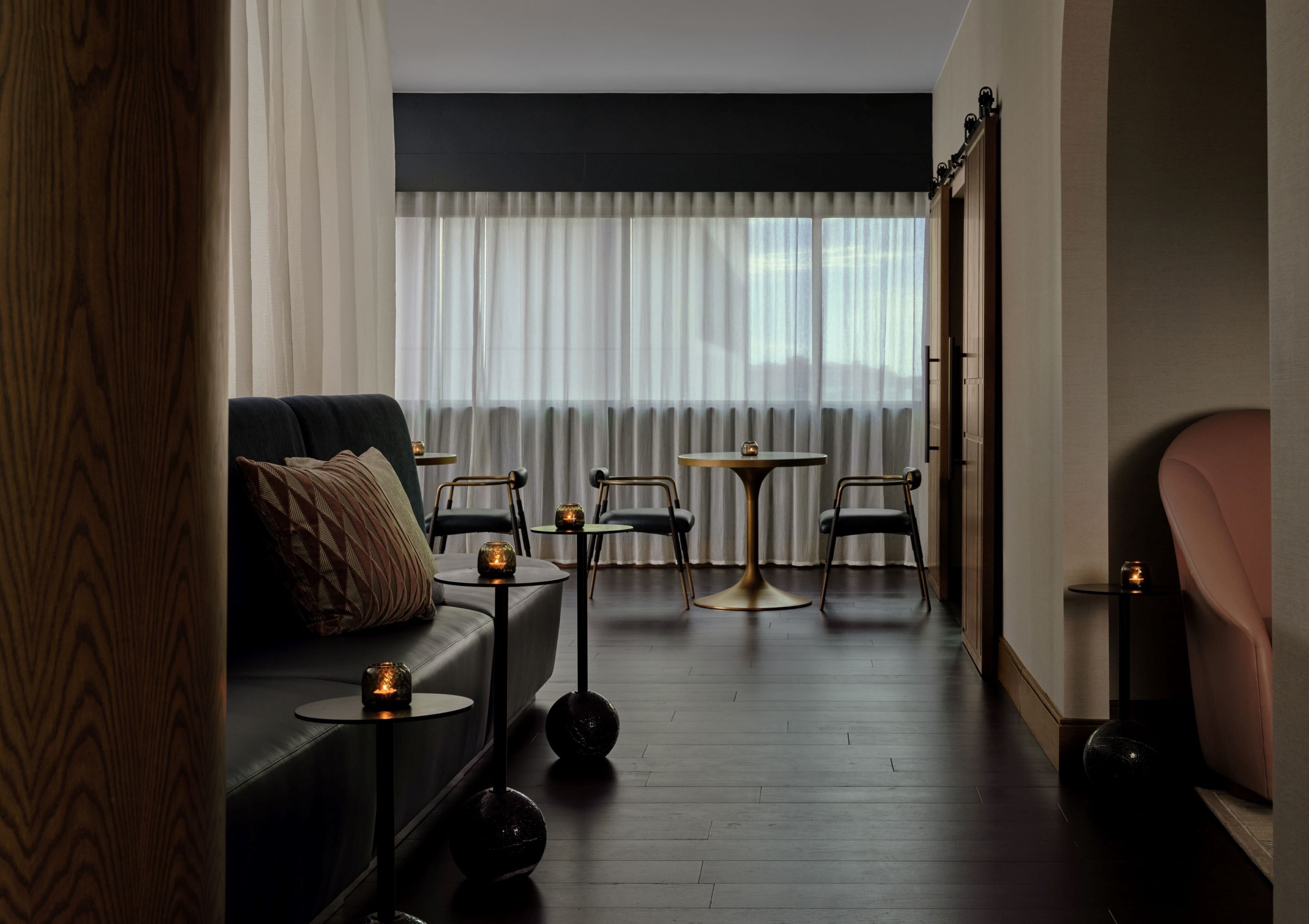 A modern hotel room with a dark wood floor, a black leather sofa, a round table with chairs, and a large window with sheer curtains.