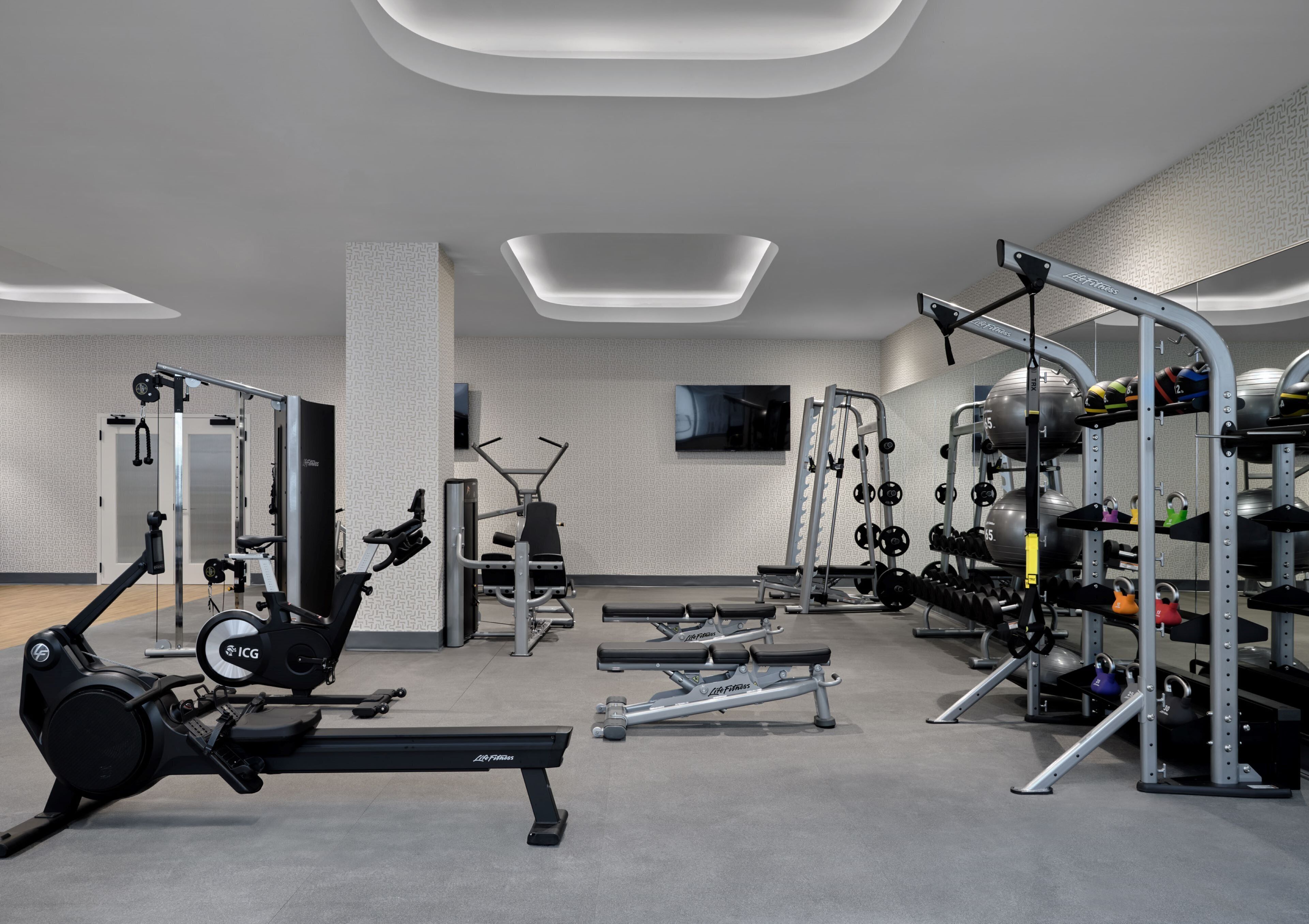 A modern gym with cardio machines, weightlifting equipment, and a TV screen on the wall.