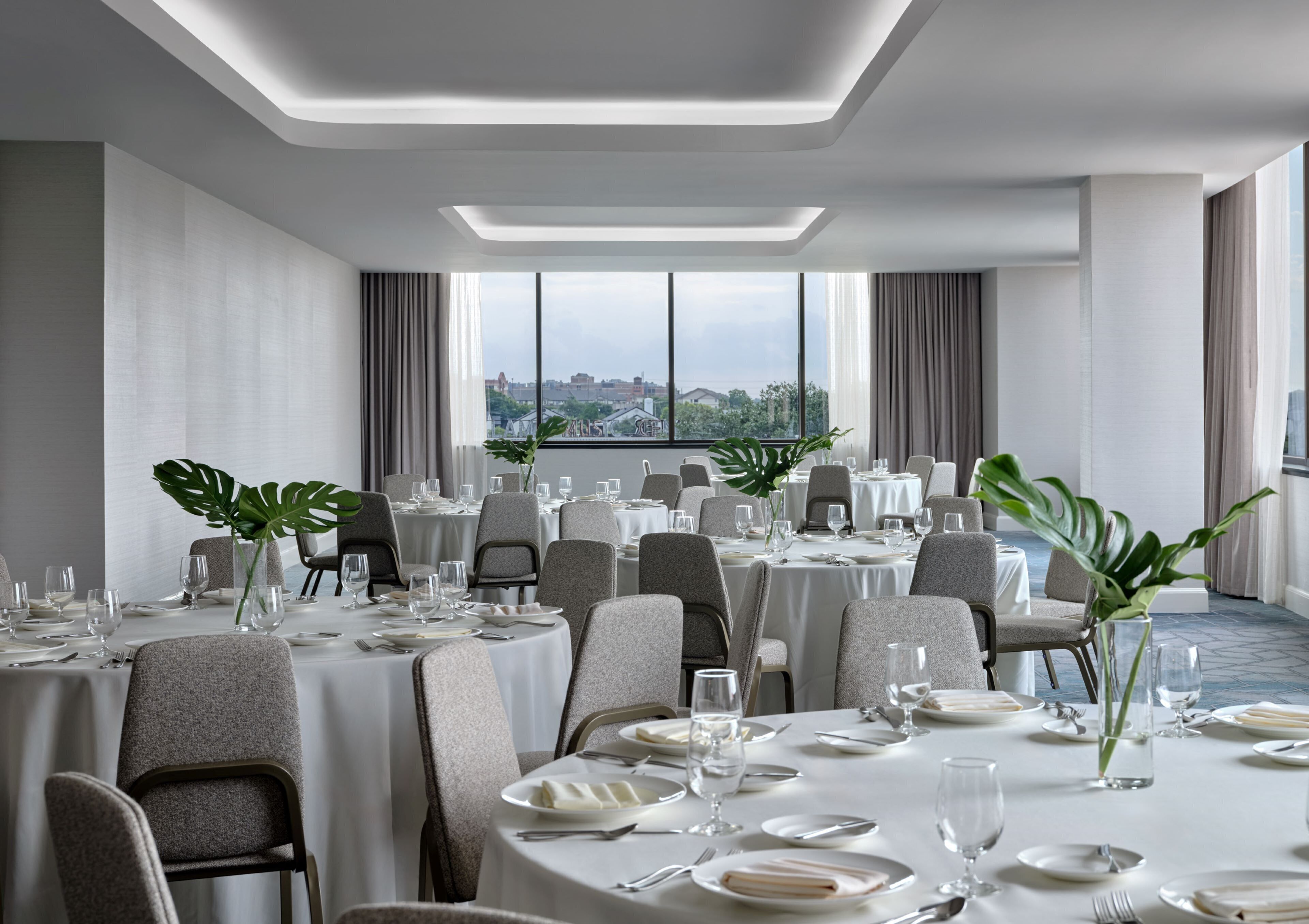 A modern dining room with round tables, grey upholstered chairs, and large windows offering a city view.