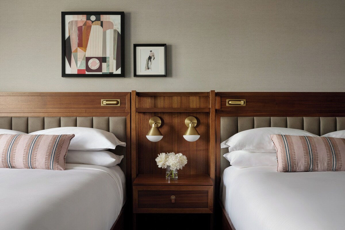 Habitación de hotel con dos camas, cabecero de madera, mesita de noche con un jarrón de flores y dos lámparas de pared.