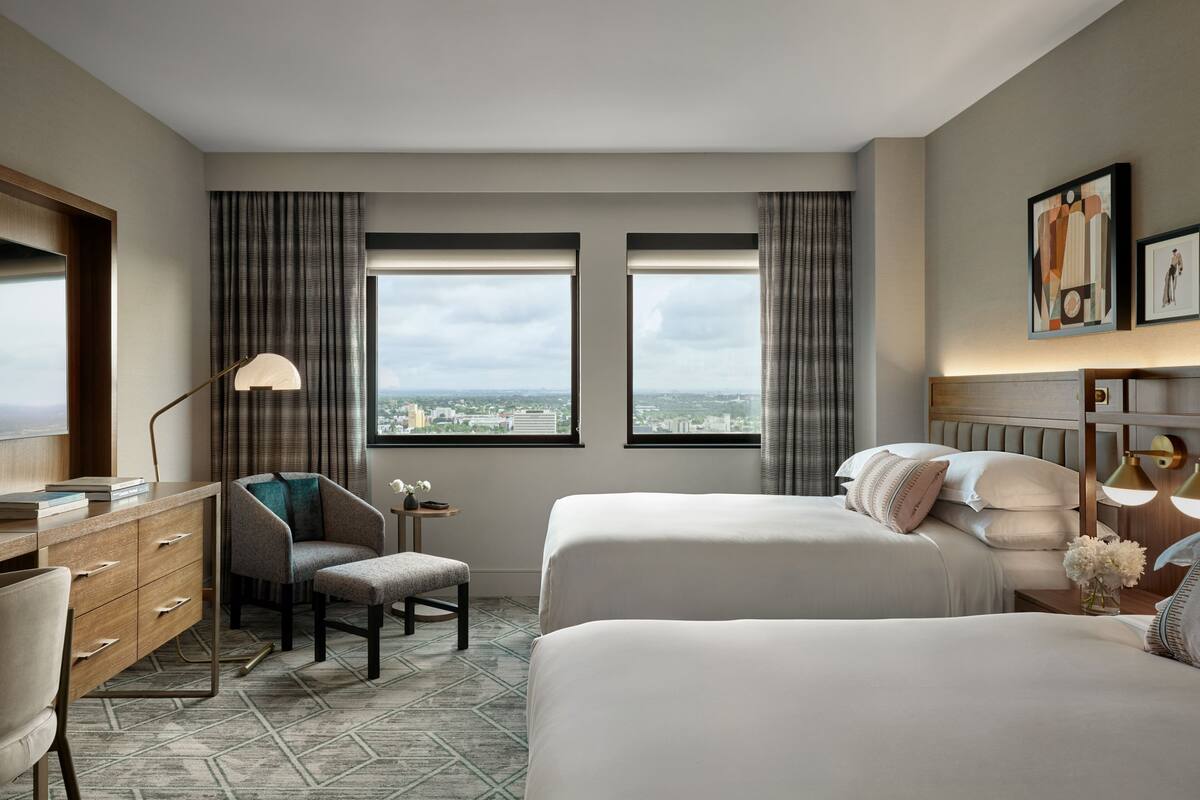 A hotel room with two beds, a desk, a chair, a lamp, and a view of the city through large windows.