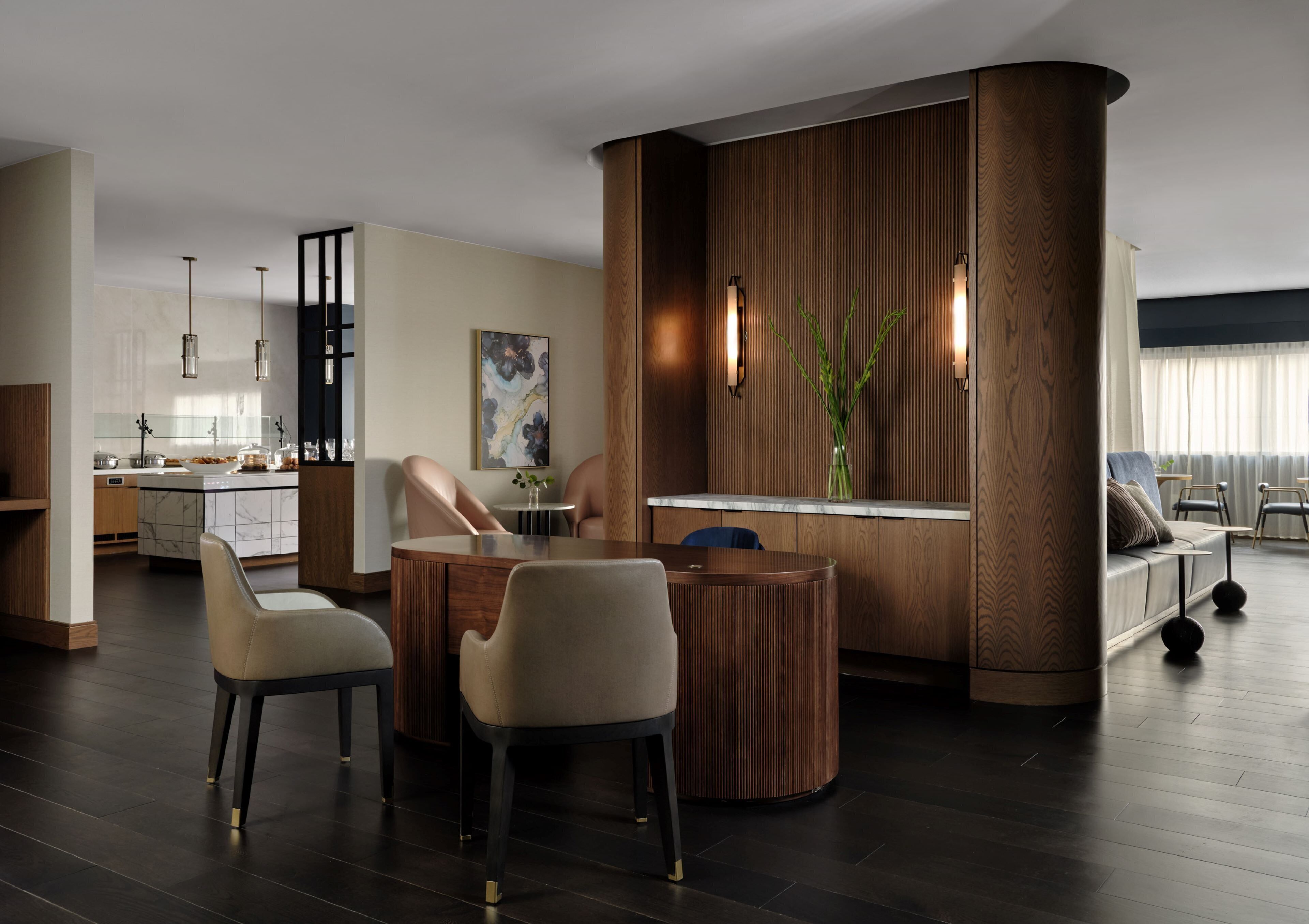 Una sala de estar moderna con una mesa de centro redonda de madera, sillas color beige, un muro de acento de madera y vistas a la cocina al fondo.