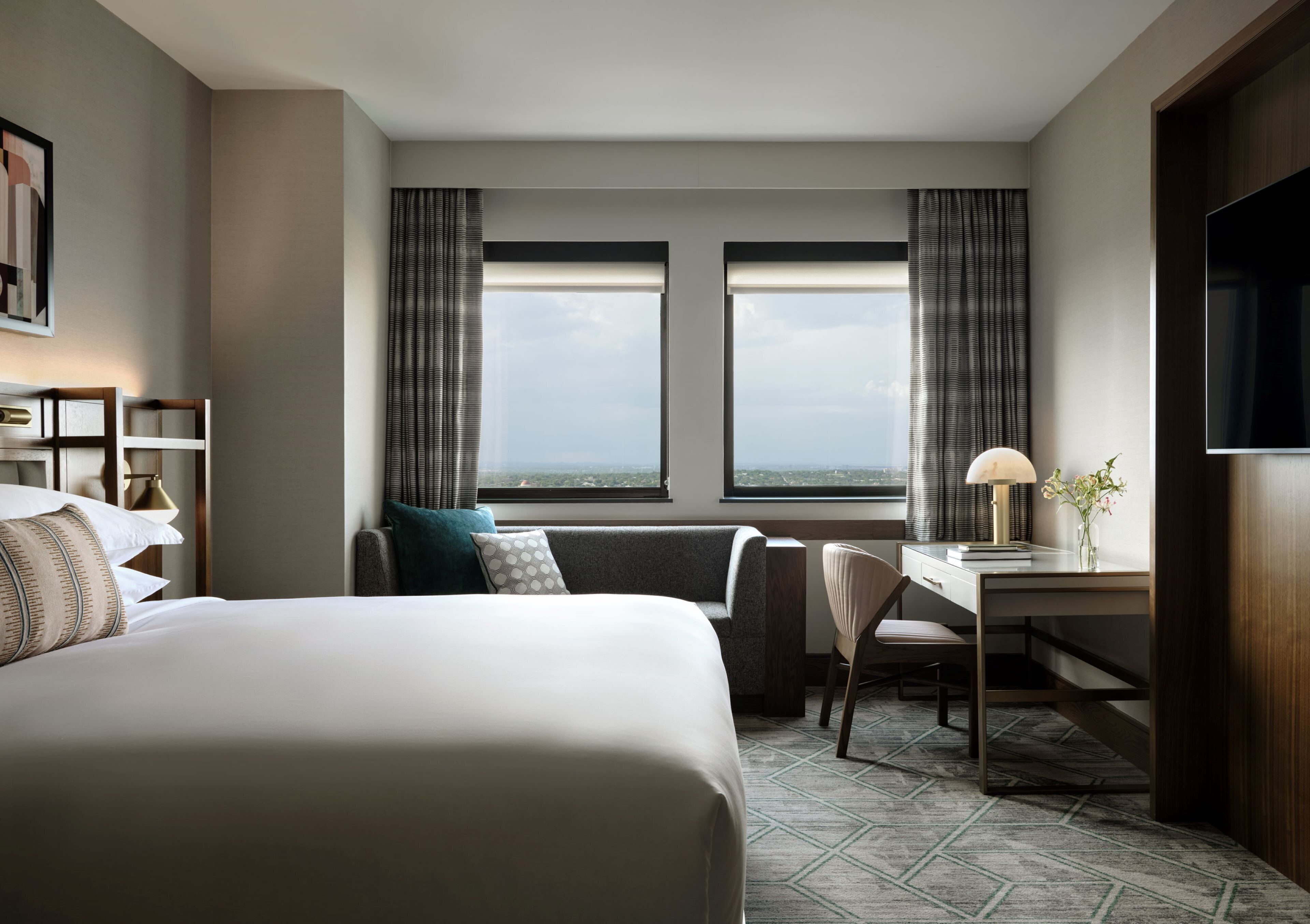 A hotel room with a large bed, a desk with a lamp, a chair, and a TV. There are two windows with curtains, and a view of the cityscape is visible through the windows.